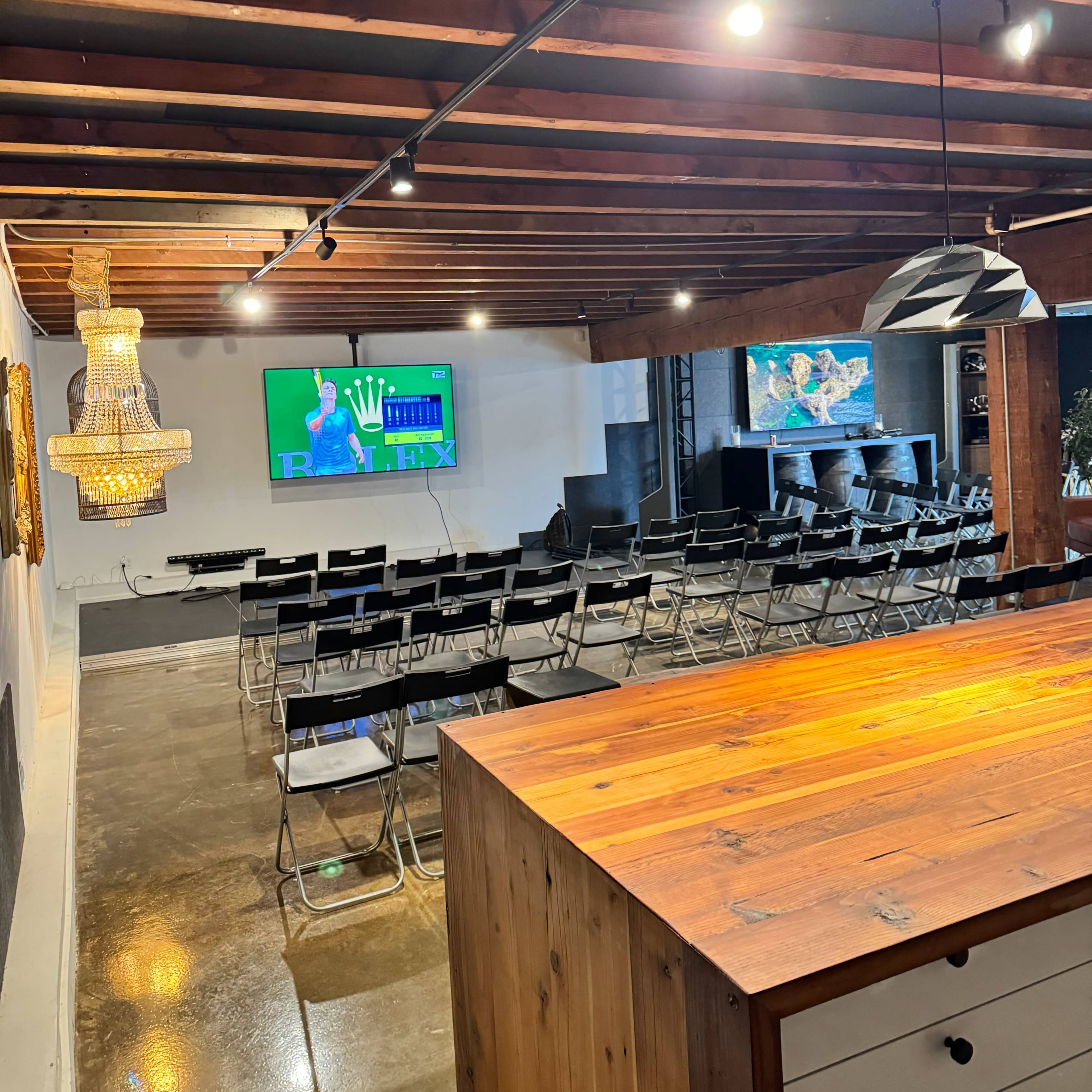 A well-lit meeting room with rows of black chairs facing a screen displaying a presentation, complemented by a wooden counter and decorative lighting in the background.