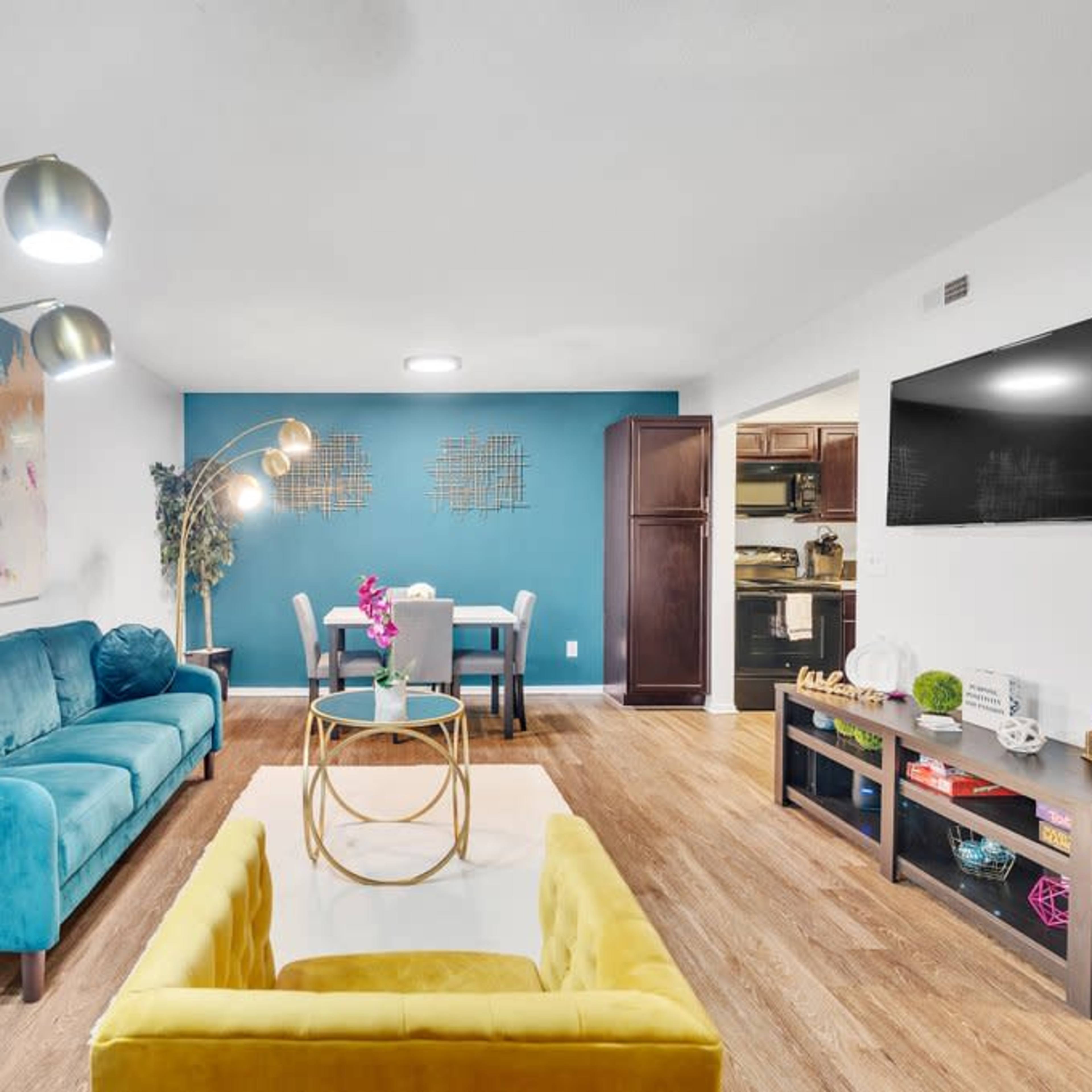 A modern living room featuring a teal sofa, a yellow armchair, a round coffee table, and a dining area with a light blue wall in the background.