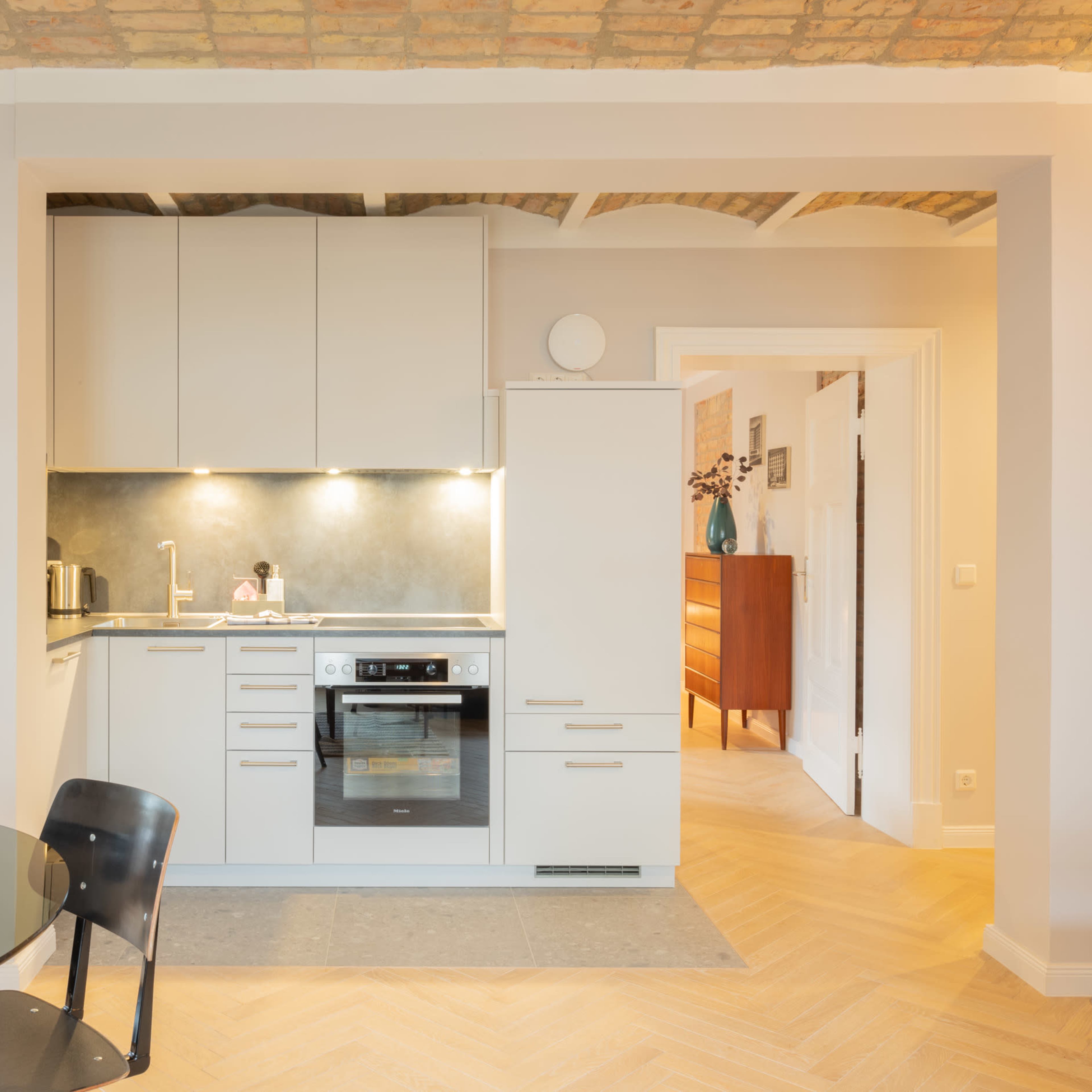 A modern kitchen adjacent to a dining area, featuring light-colored cabinetry, a stove, and a glass dining table with black chairs.