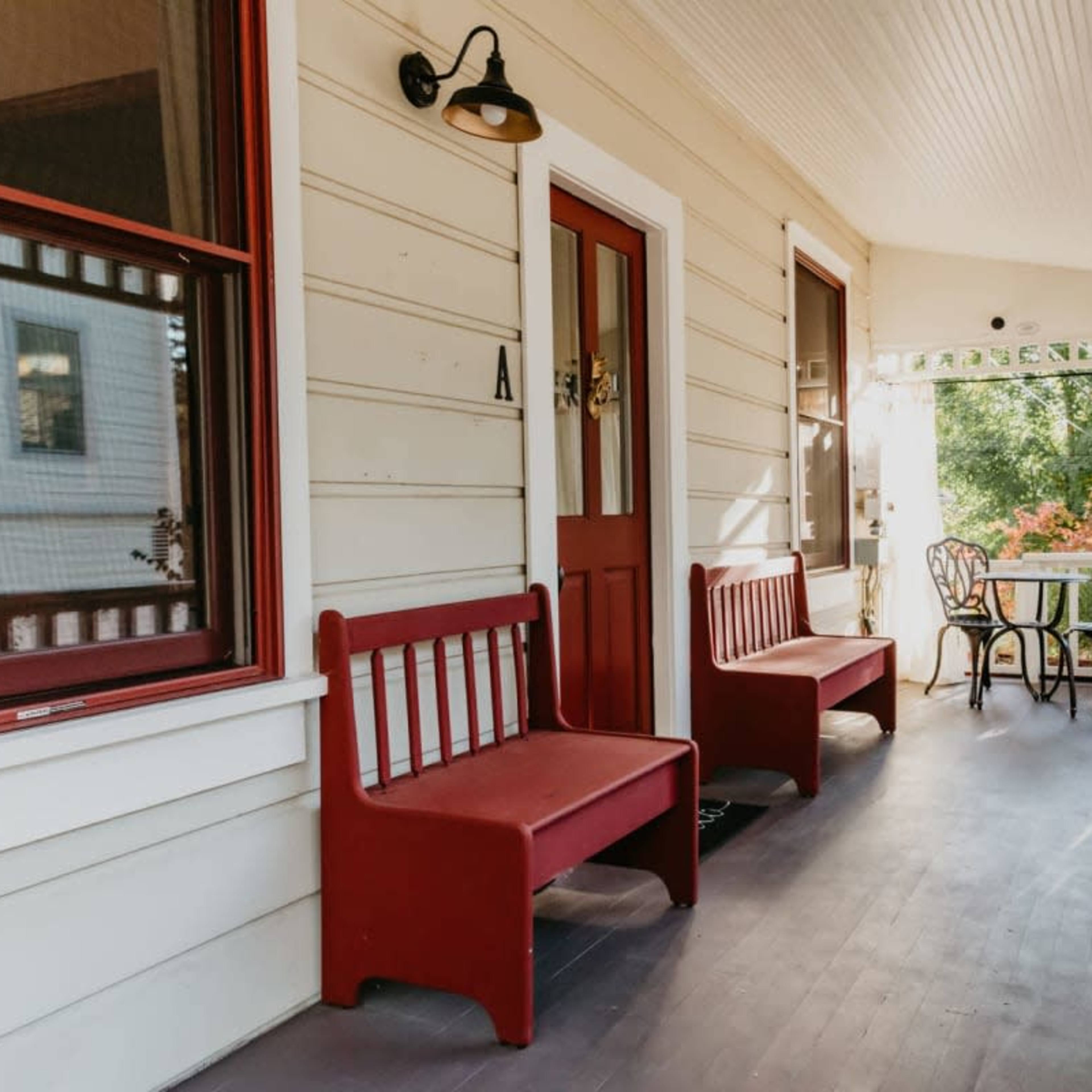 A porch with two red benches and a small table with chairs, surrounded by greenery.