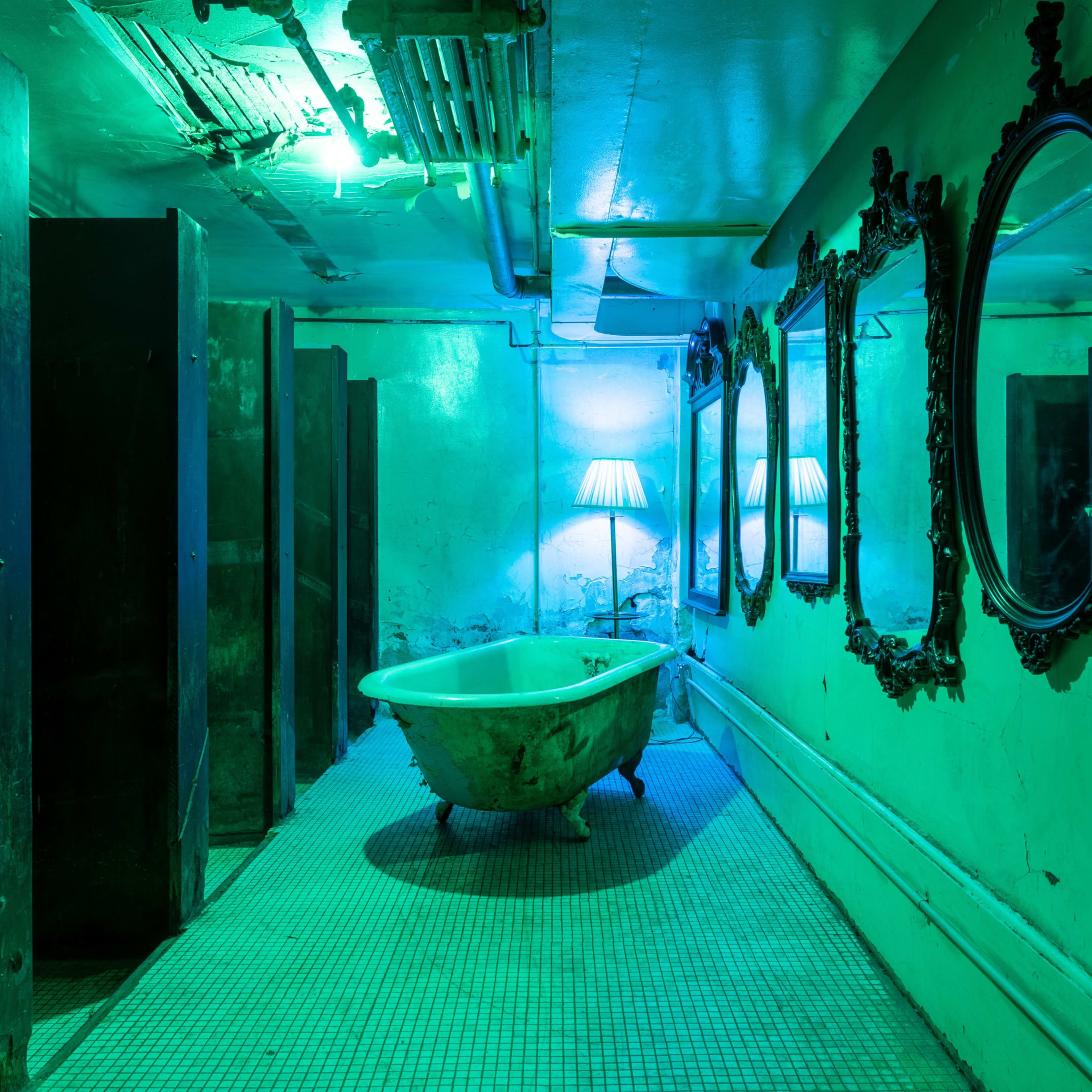 A dimly lit restroom with a green hue, featuring a vintage bathtub, multiple mirrors, and a lamp in the corner.