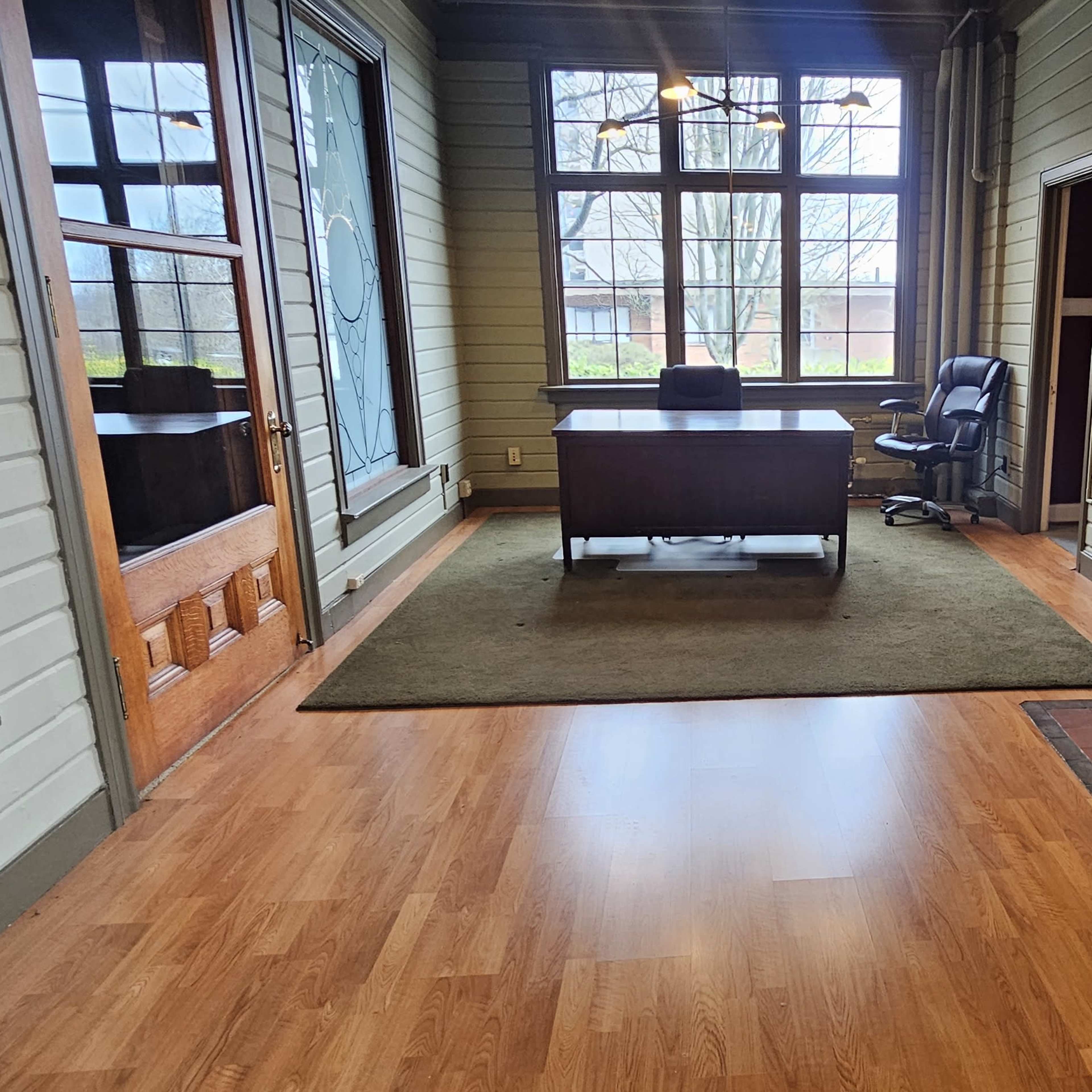 An office space featuring a wooden desk with a chair, a fireplace, and large windows allowing natural light to enter.