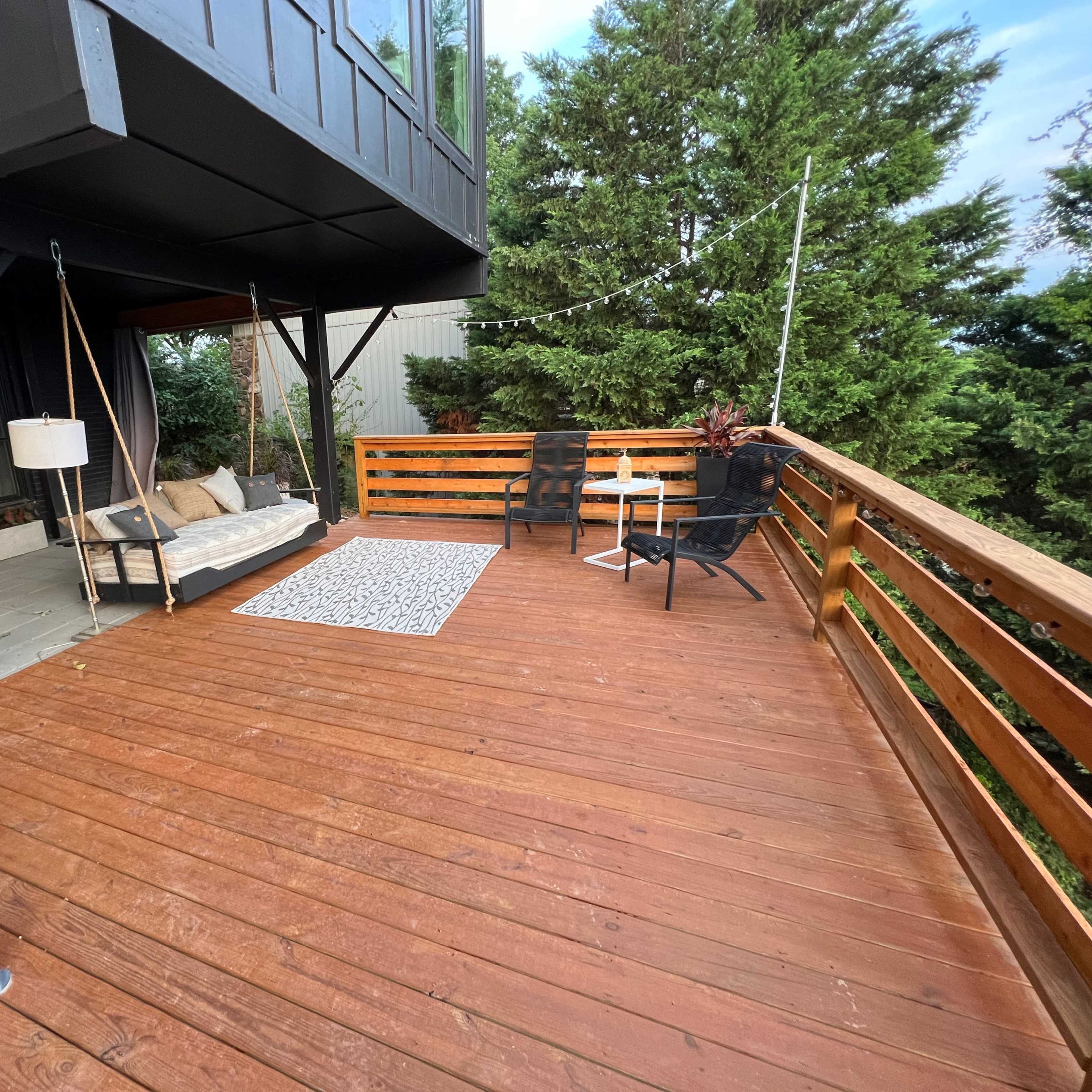 A wooden deck with seating, a rug, and greenery in the background.