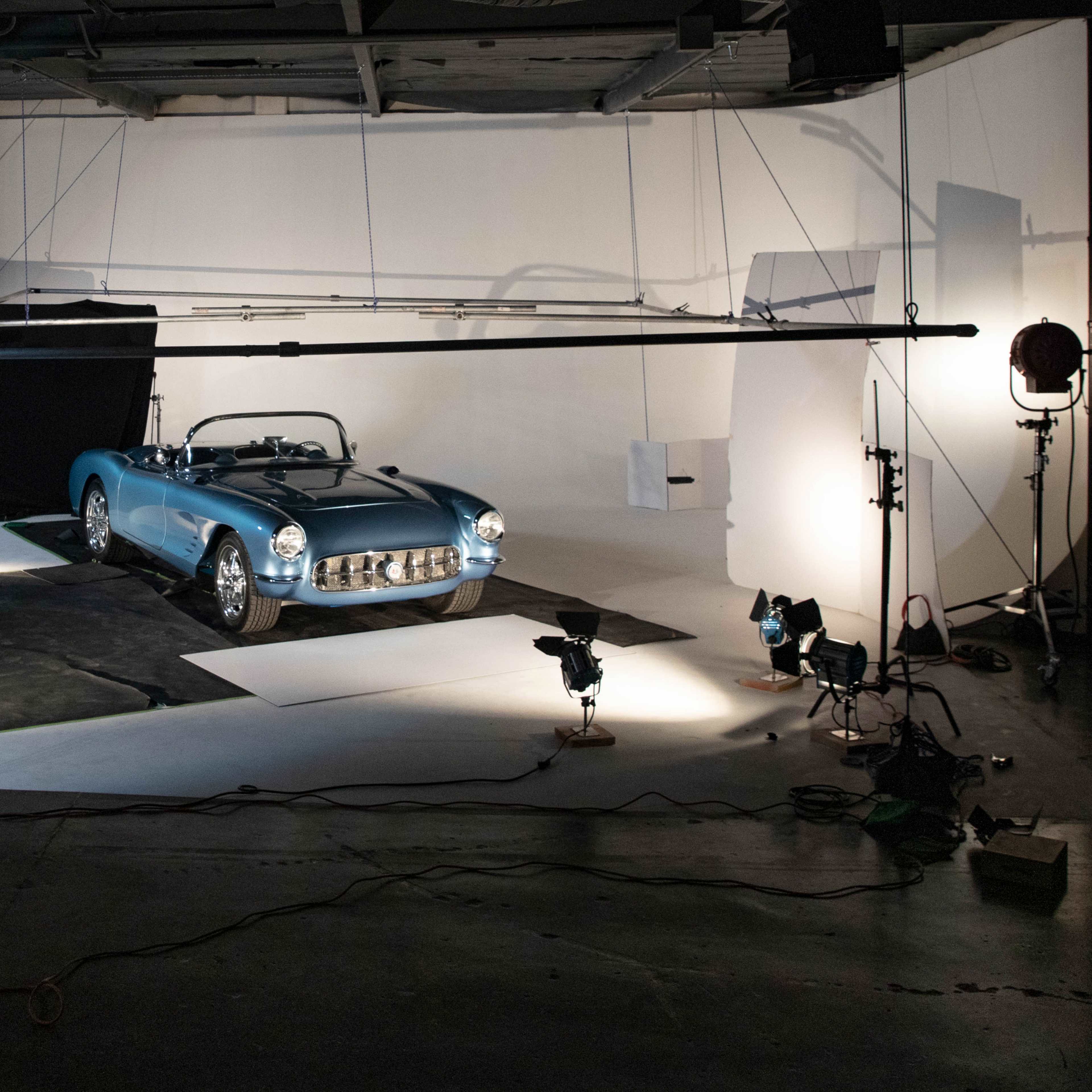 A classic blue convertible car is positioned on a photo studio set, surrounded by lighting equipment and a ladder.