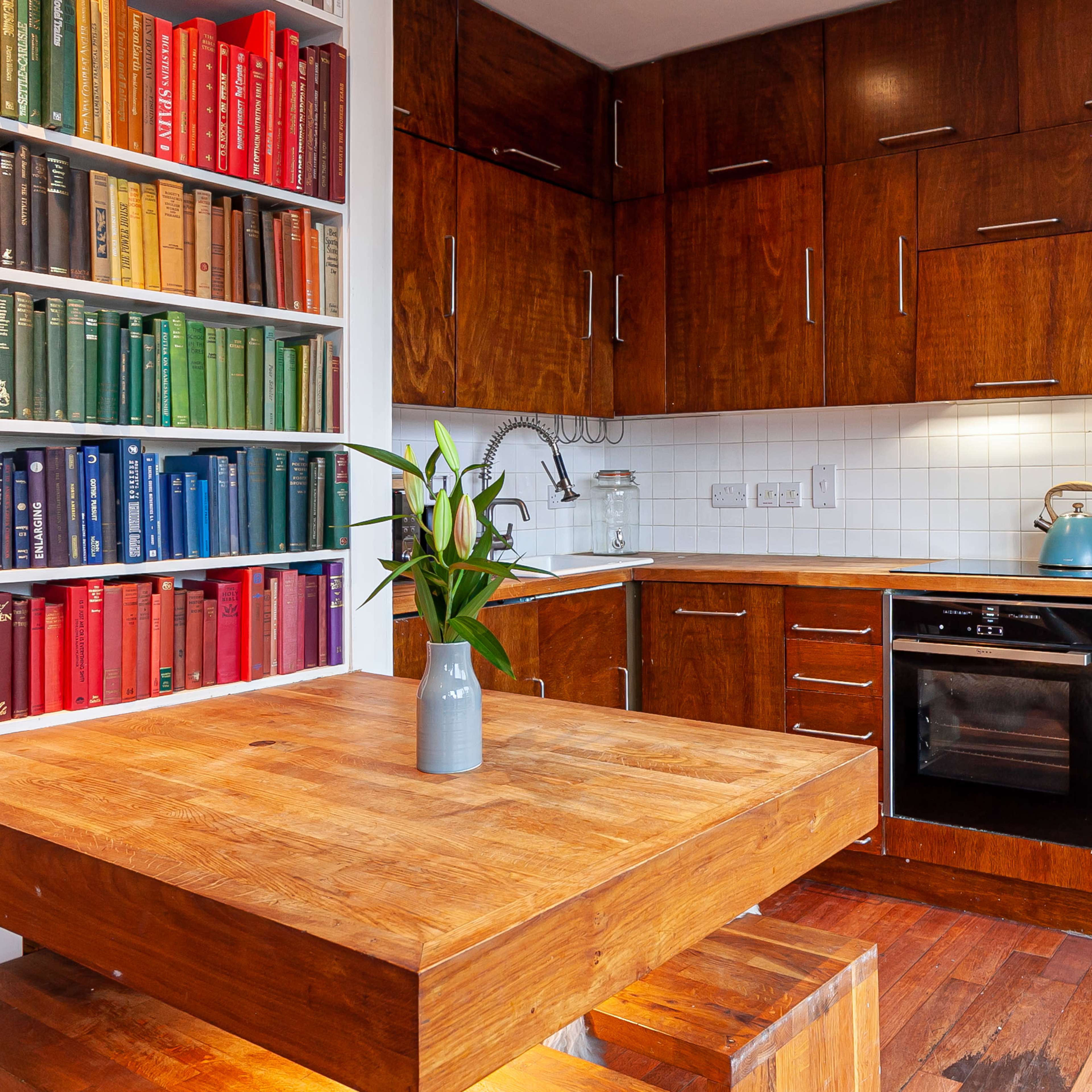 A modern kitchen with wooden cabinets, a colorful bookshelf filled with books, and a wooden dining table with a small vase of flowers.