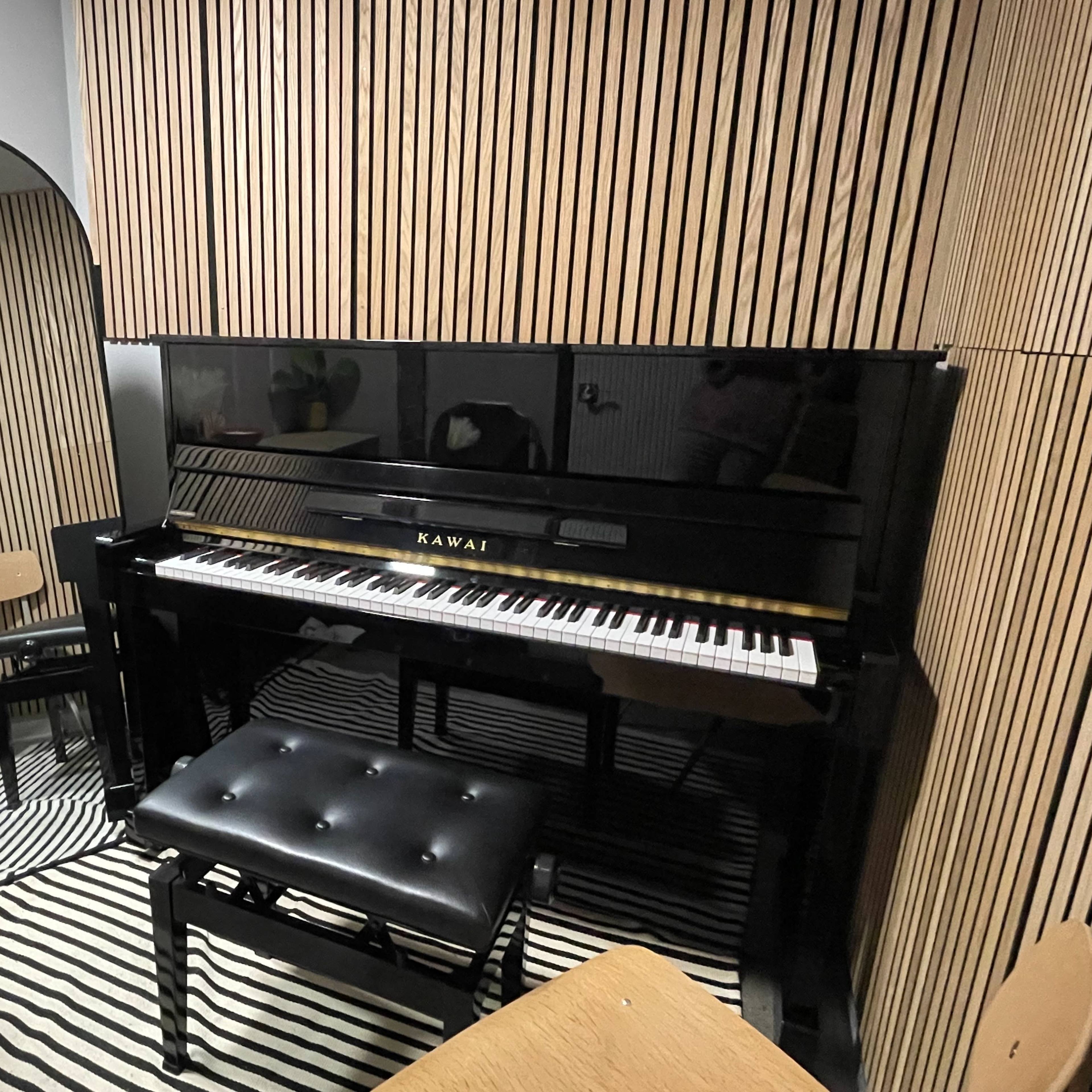 A Kawai piano in a room with vertical wooden paneling, accompanied by a mirrored wall and a small seating area.