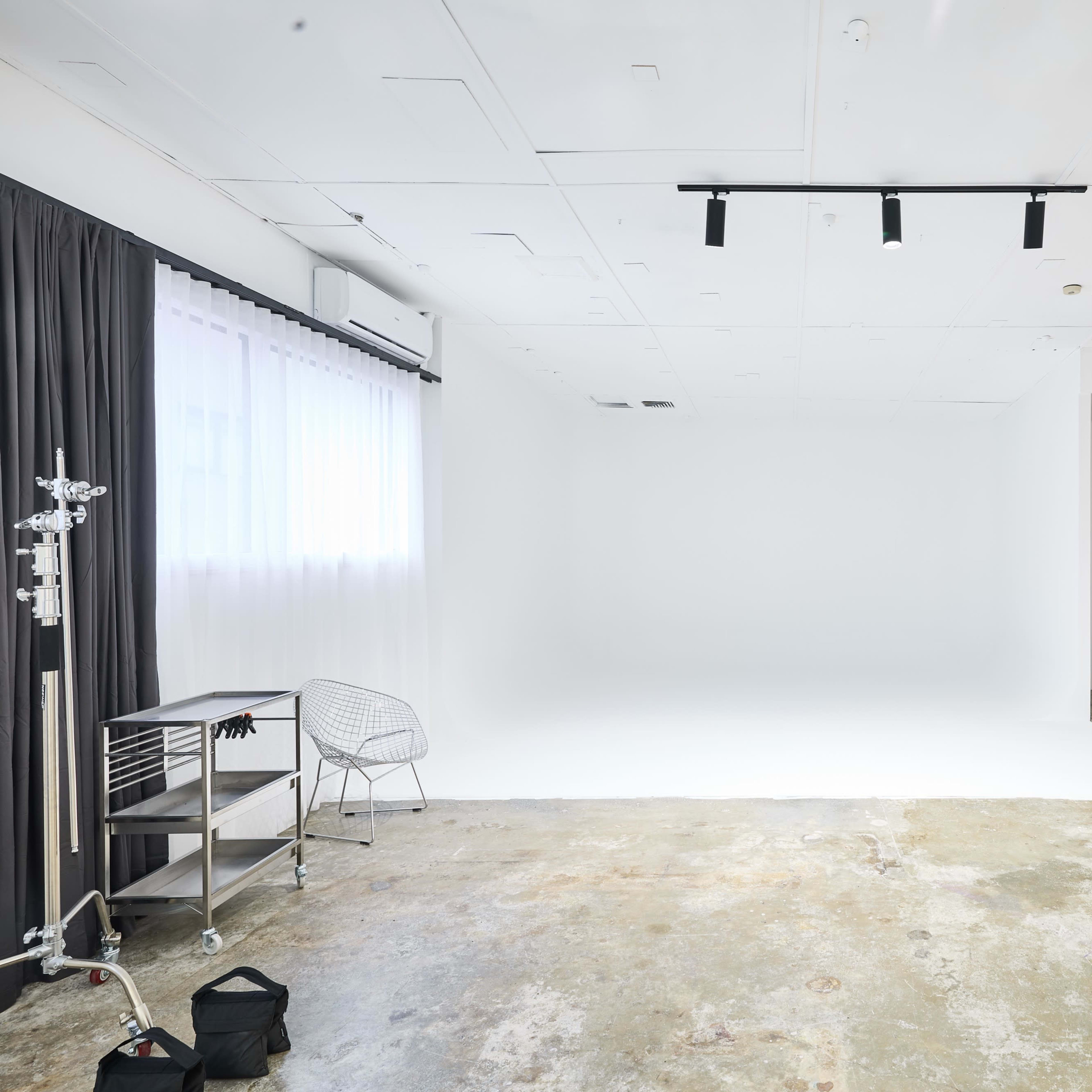 A spacious, well-lit studio features a white backdrop, concrete flooring, and minimal furnishings including a chair and a large mirror.