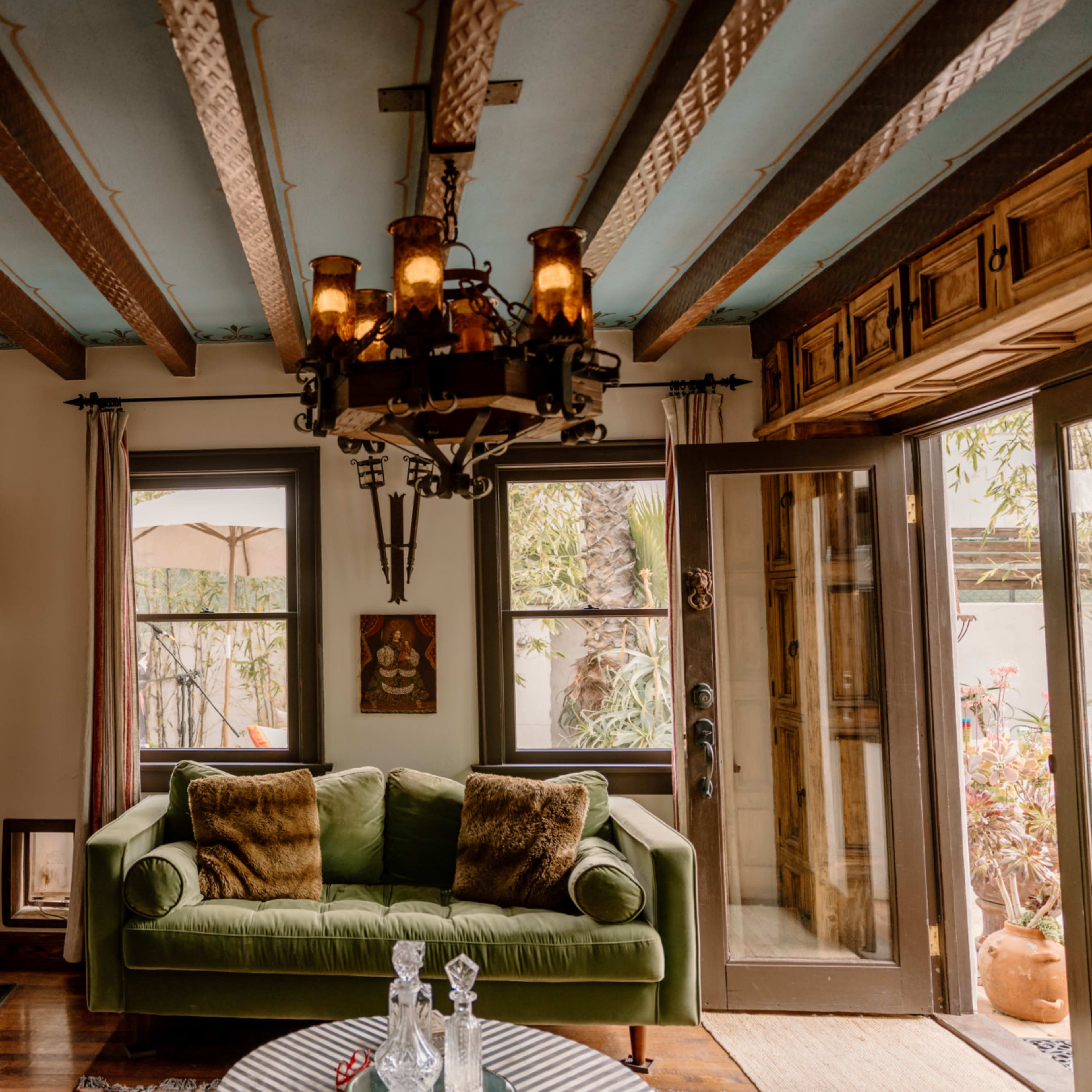 A cozy living room features a green couch, wooden beams overhead, and a chandelier, with large windows providing a view of an outdoor area.