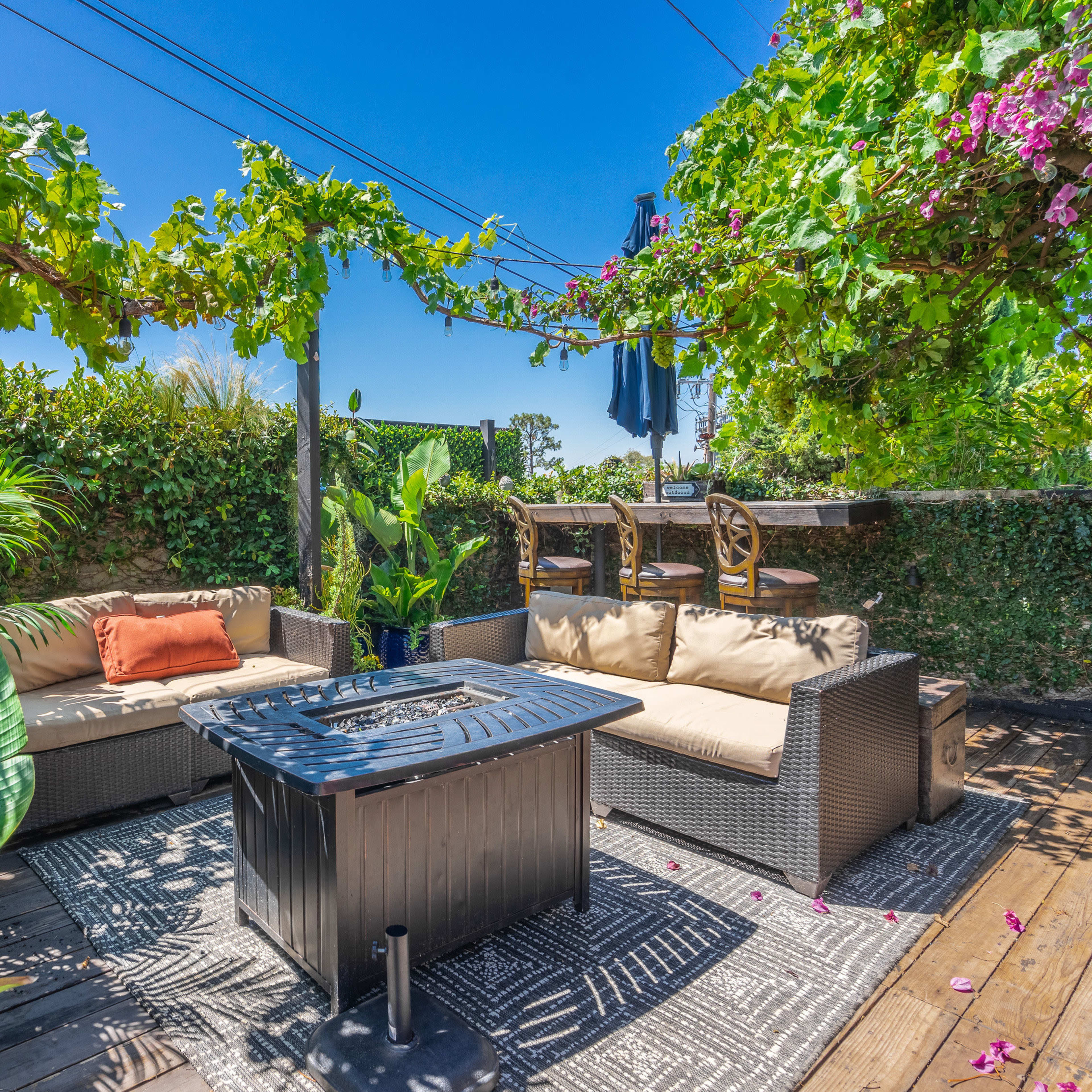 A patio area featuring a seating arrangement with cushions, a fire pit, and lush greenery overhead.