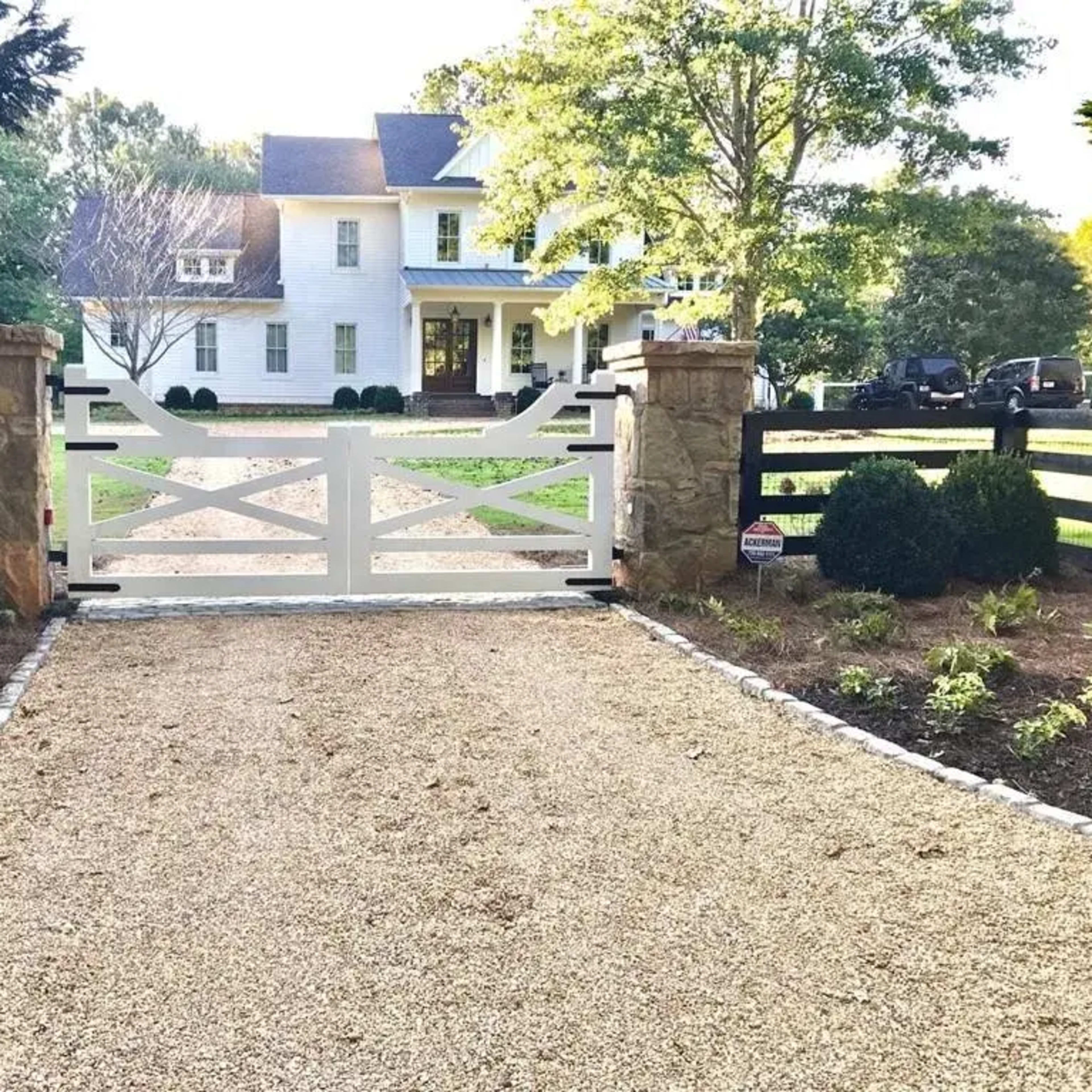 A white gate with stone pillars stands at the entrance of a gravel driveway leading to a large white house surrounded by trees and landscaping.