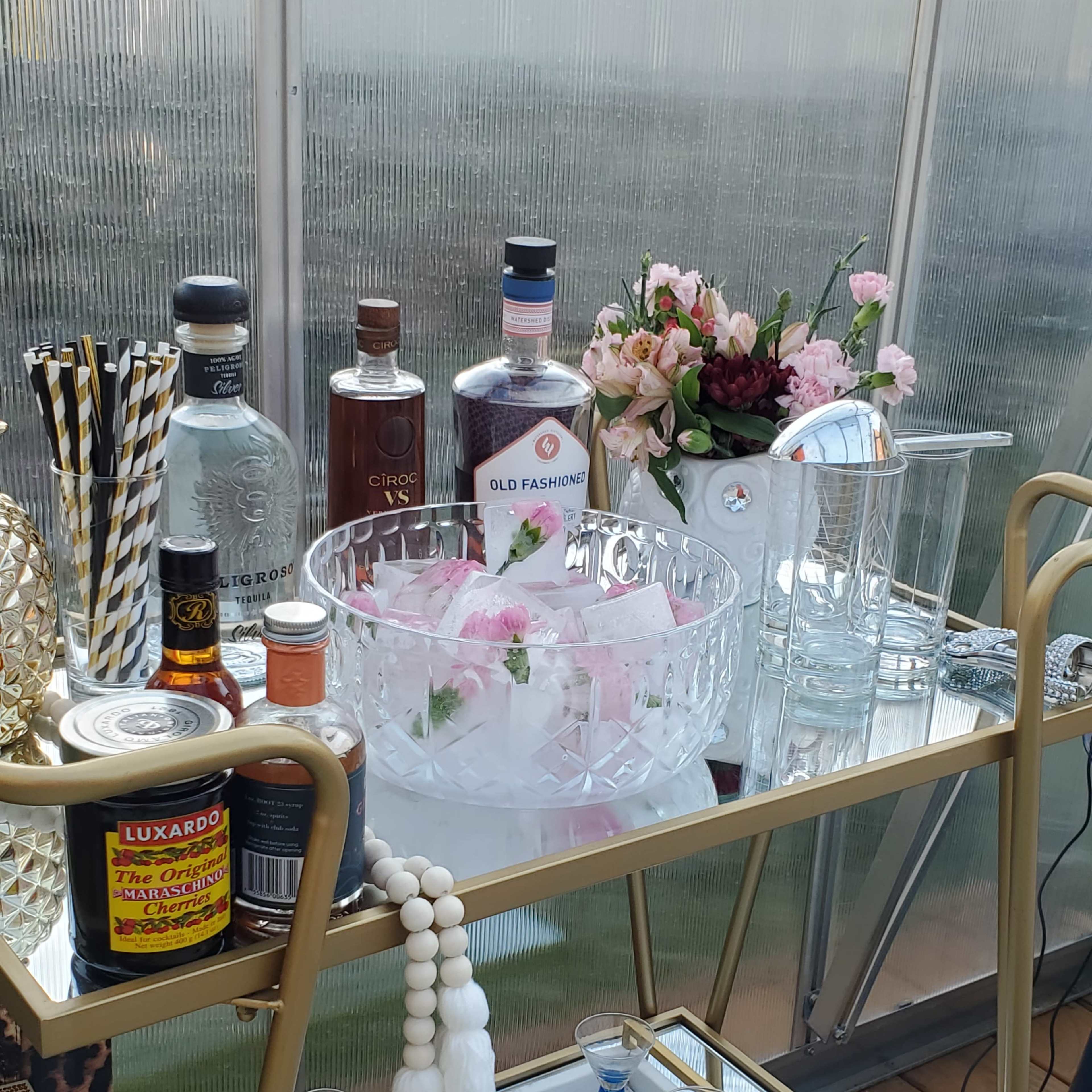 A bar cart displays an array of spirits, cocktail ingredients, decorative flowers, and glassware.