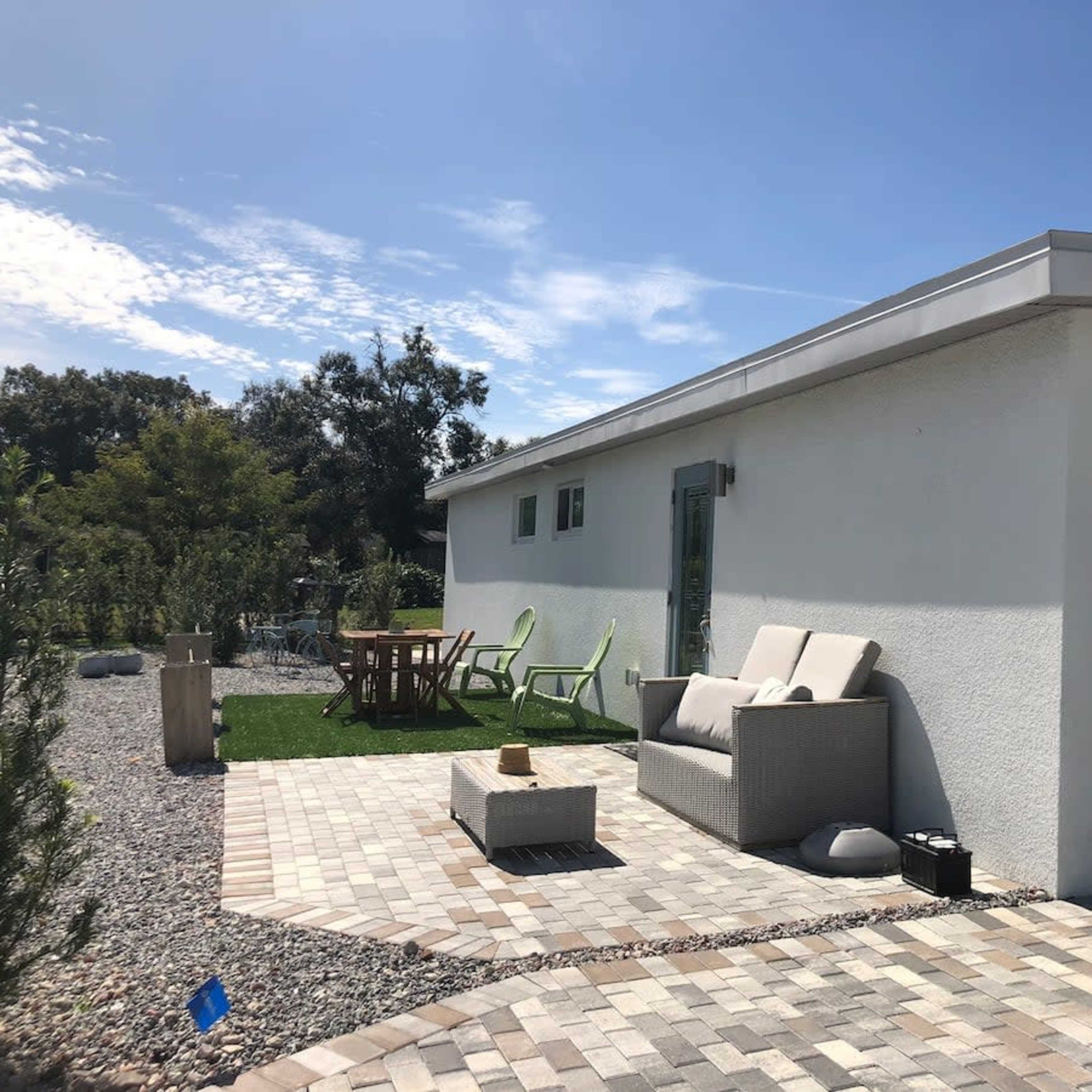 A modern house with a landscaped patio features a seating area, a dining table, and stone pathways under a clear blue sky.