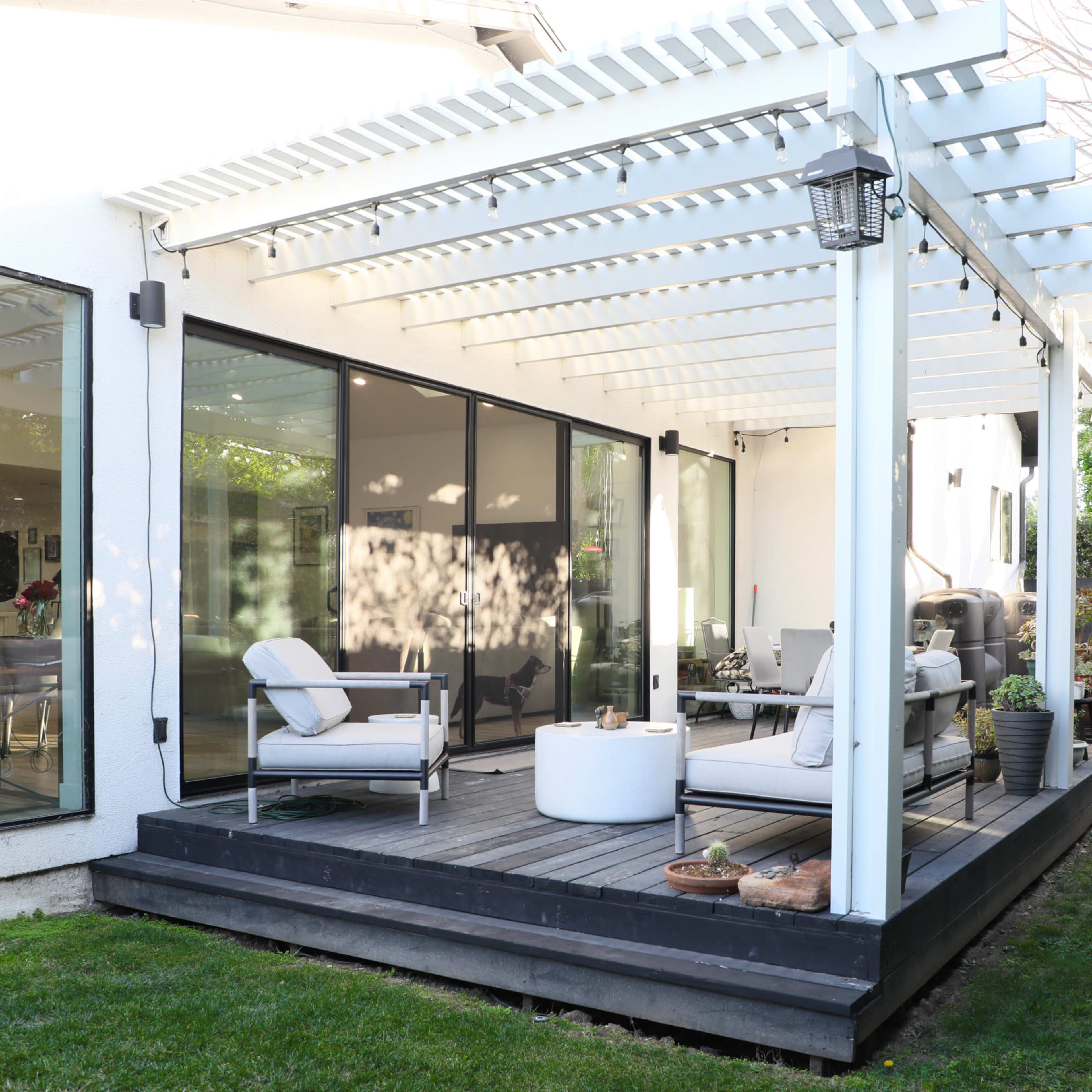 A modern patio with a covered seating area, featuring a white pergola and a mix of furniture on a wooden deck surrounded by green grass.