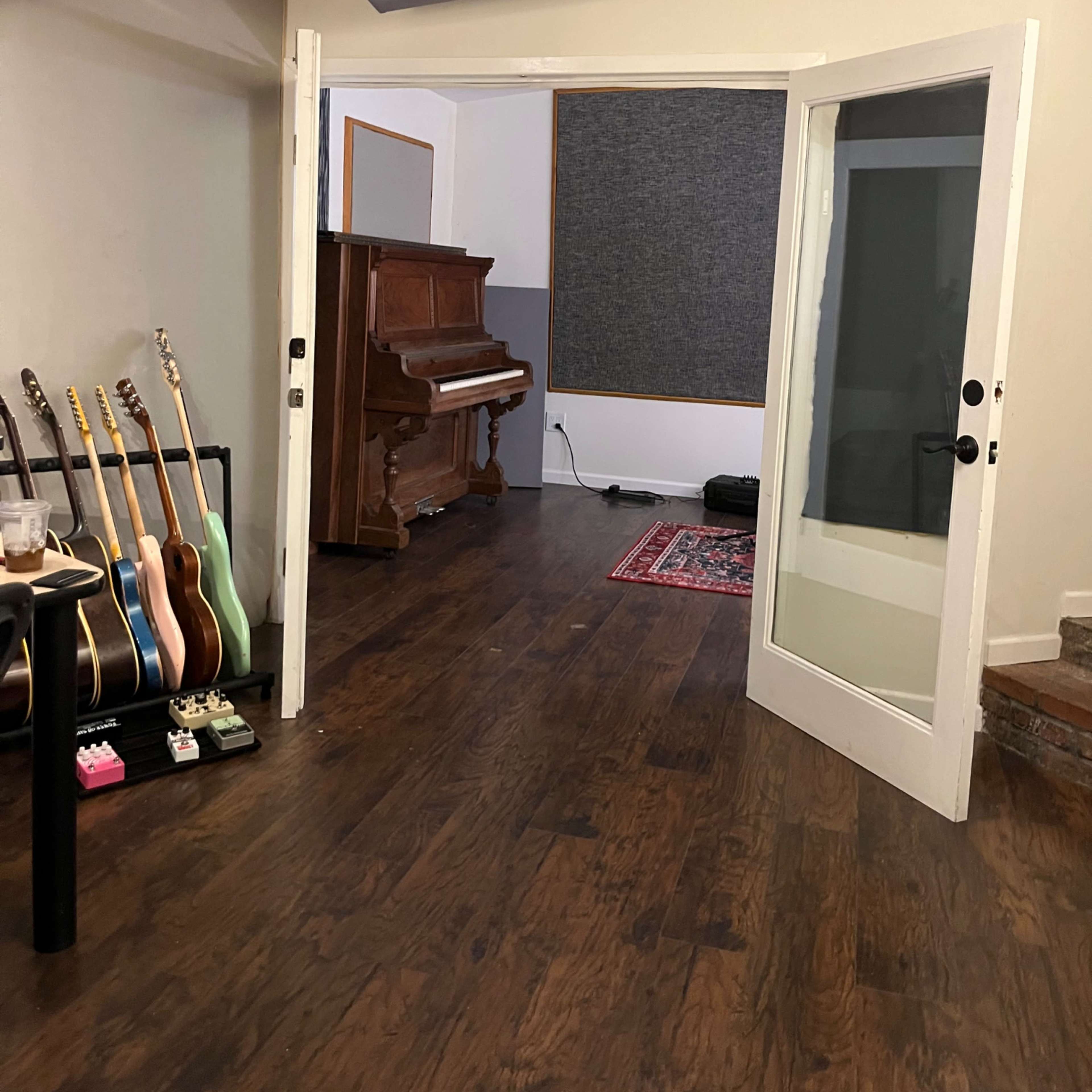 A room with an open doorway leading to a space that contains a vintage piano, several guitars on stands, and sound-absorbing panels on the walls.