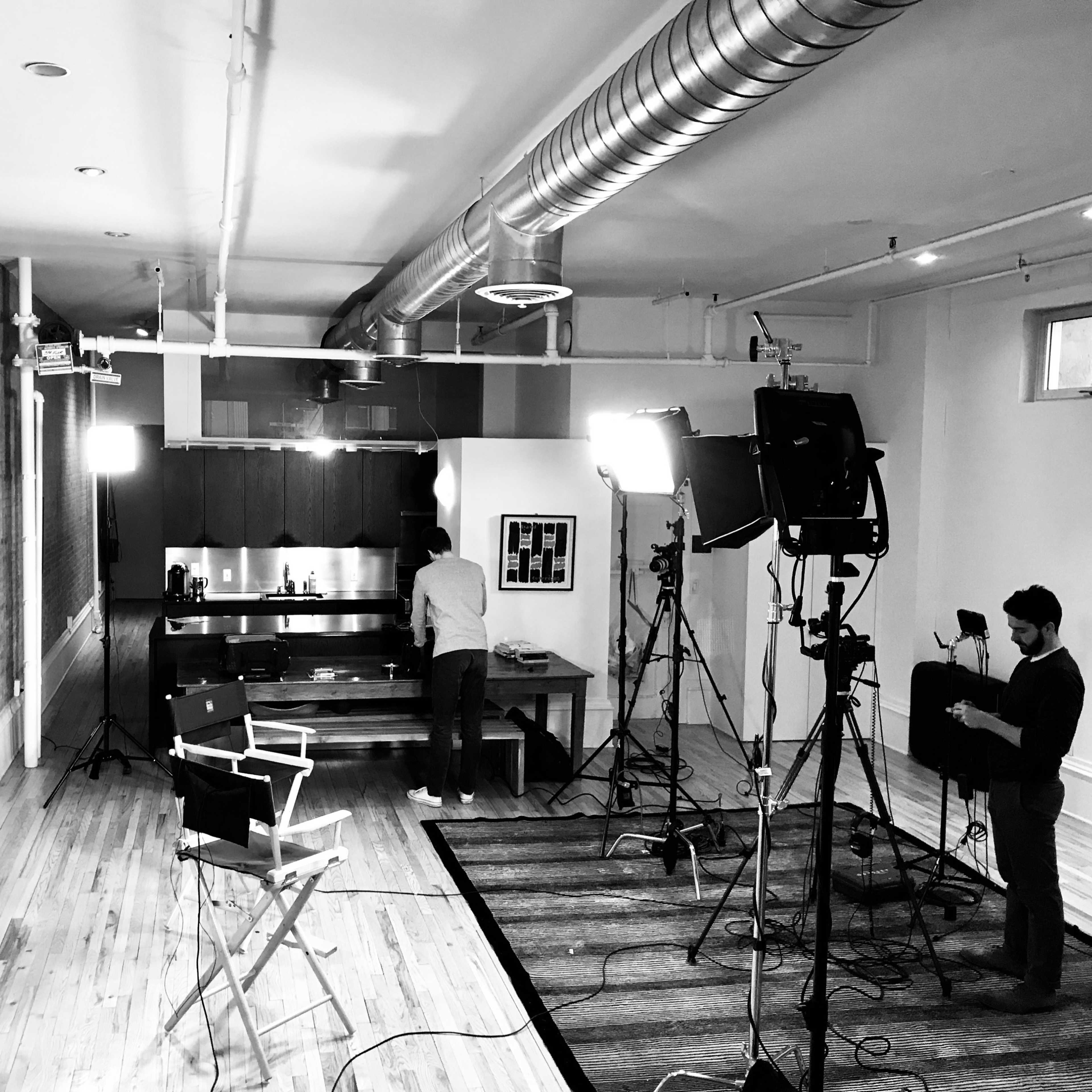 A spacious kitchen area is set up for a video shoot, with multiple cameras and lights positioned around a central wooden table where a person is preparing for filming.
