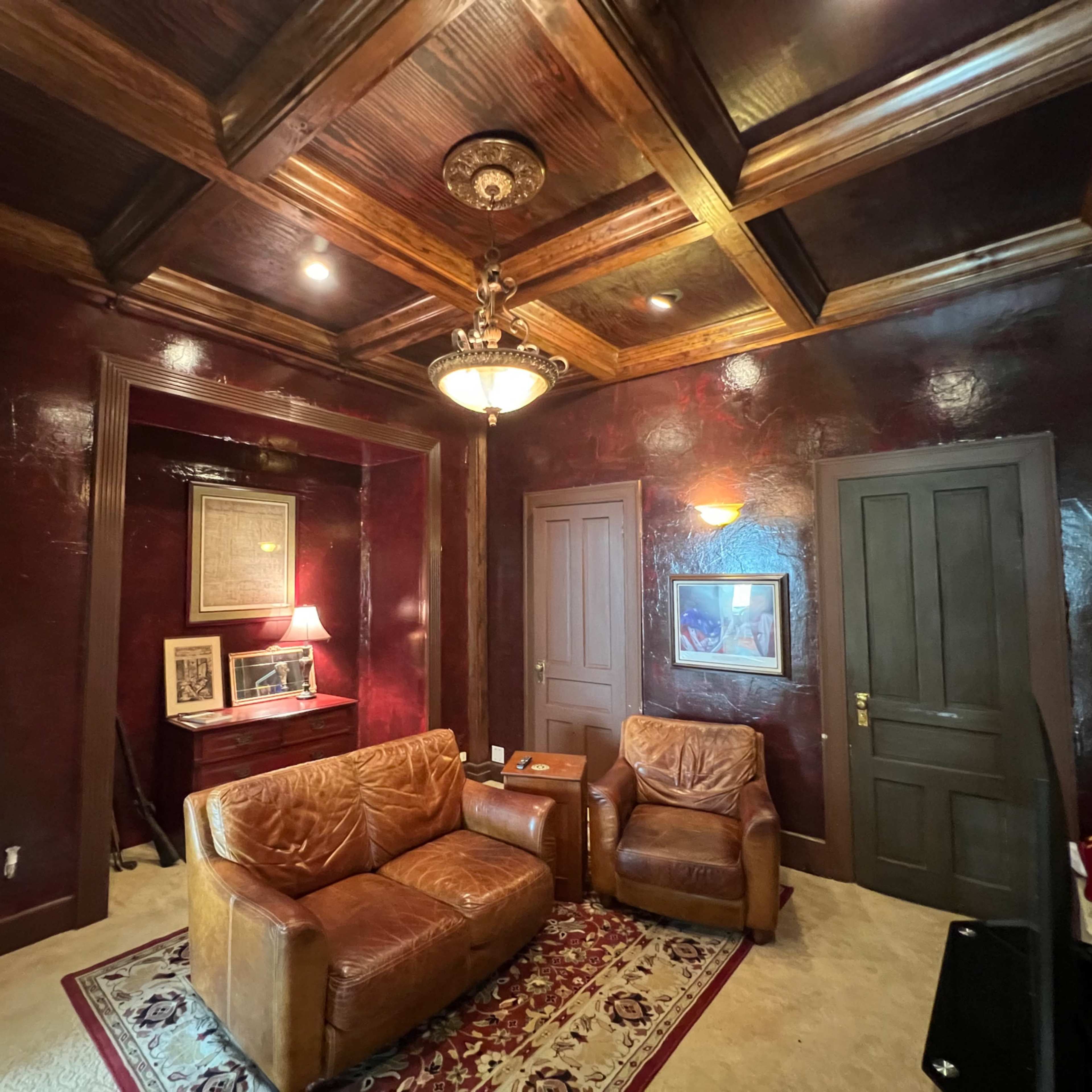 A cozy office space with dark red walls, a wooden coffered ceiling, two brown leather armchairs, and a desk with a lamp.