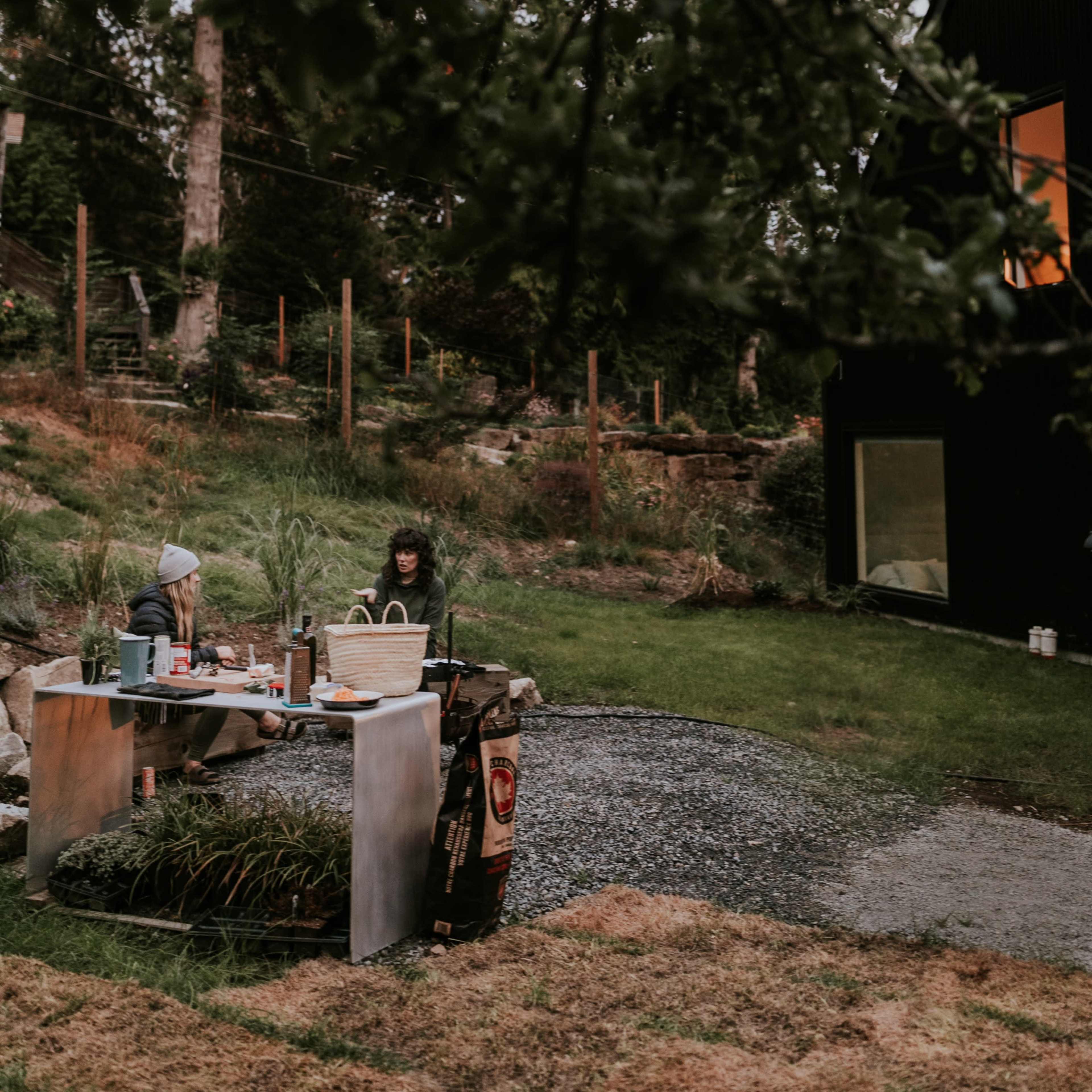 Two people are seated at a metal table in a grassy area, working amidst a backdrop of trees and a dark house.