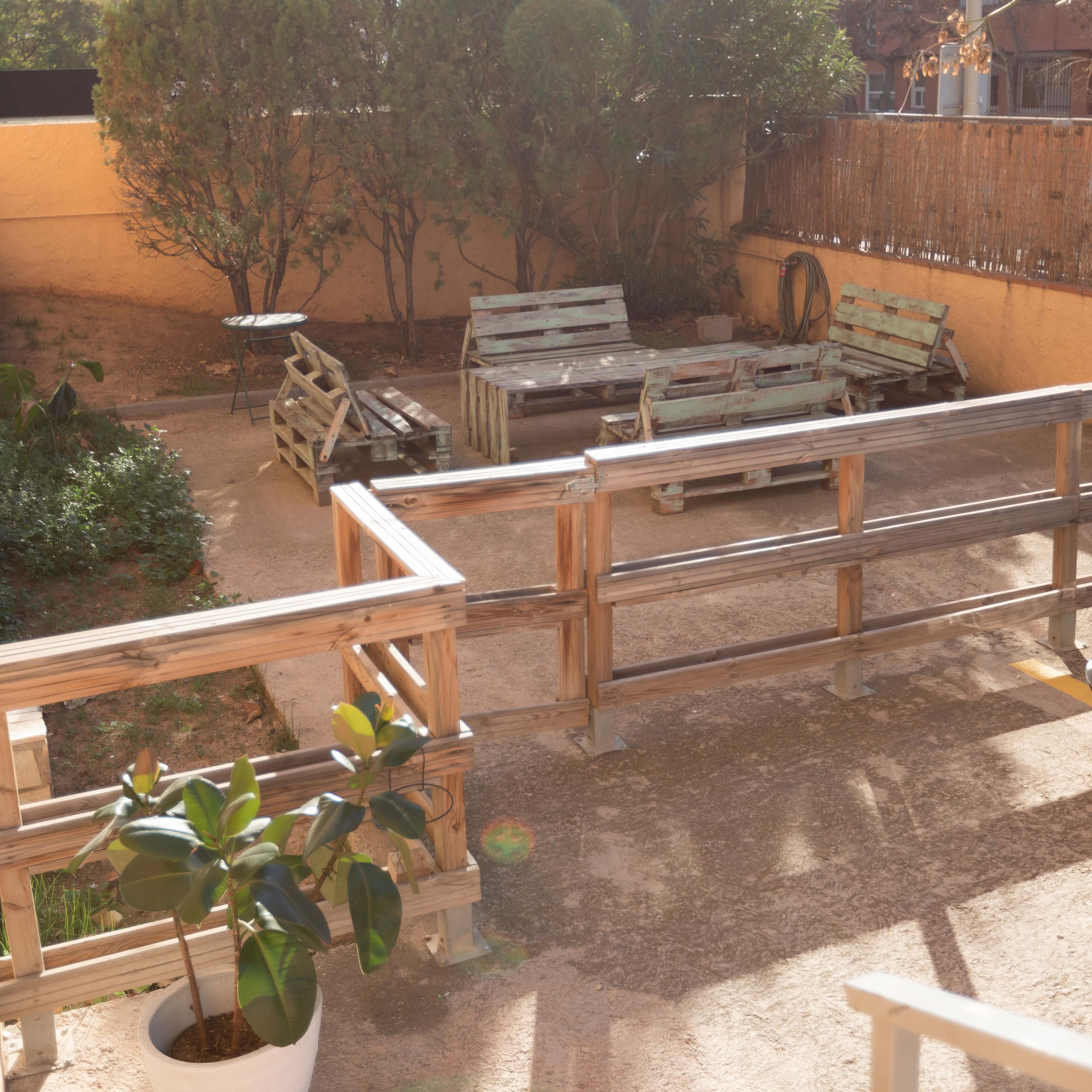 A sunlit backyard with wooden seating arrangements, a table, and a few potted plants surrounded by shrubs.