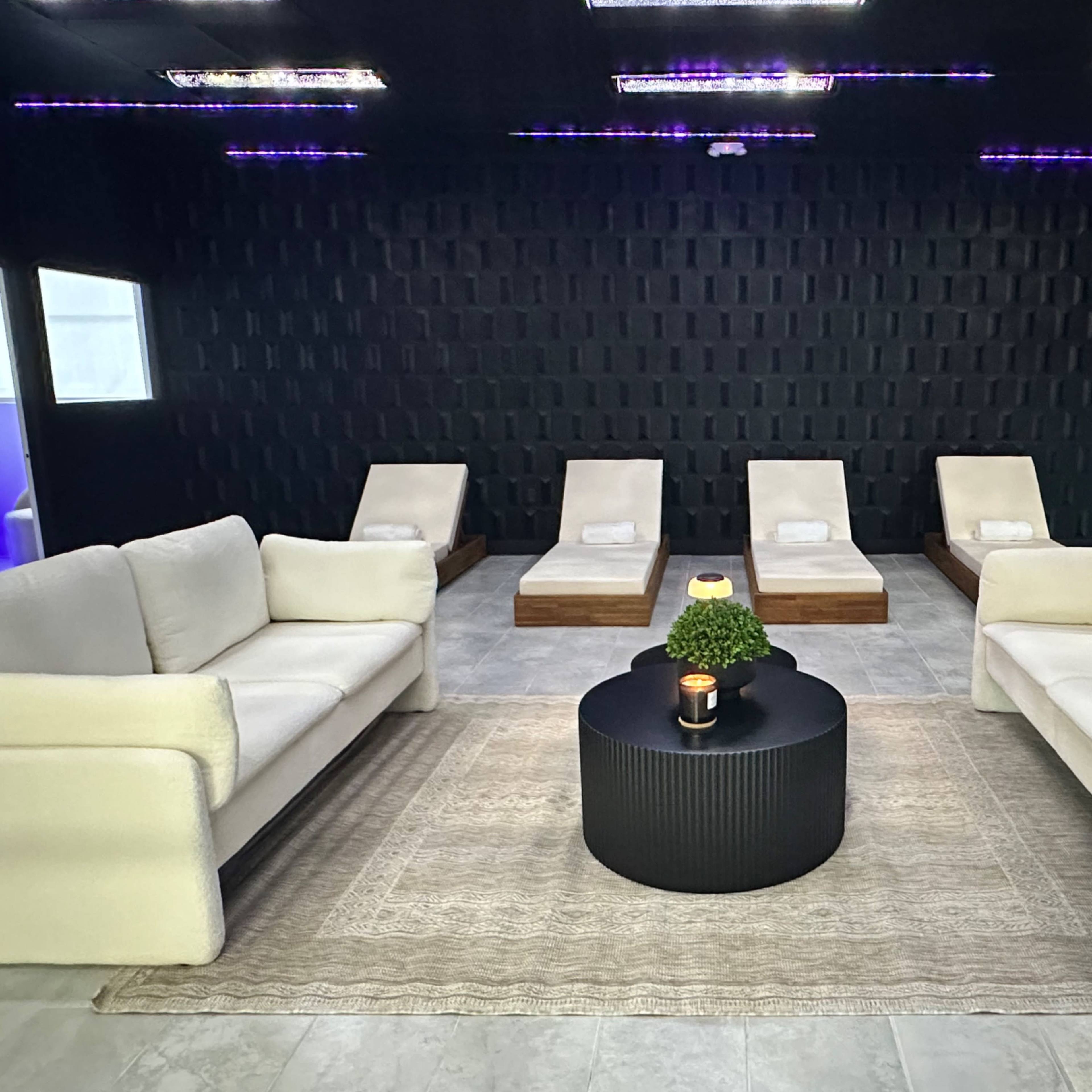 A modern lounge area with white sofas facing a round black coffee table, surrounded by wooden lounge chairs and a dark textured wall.