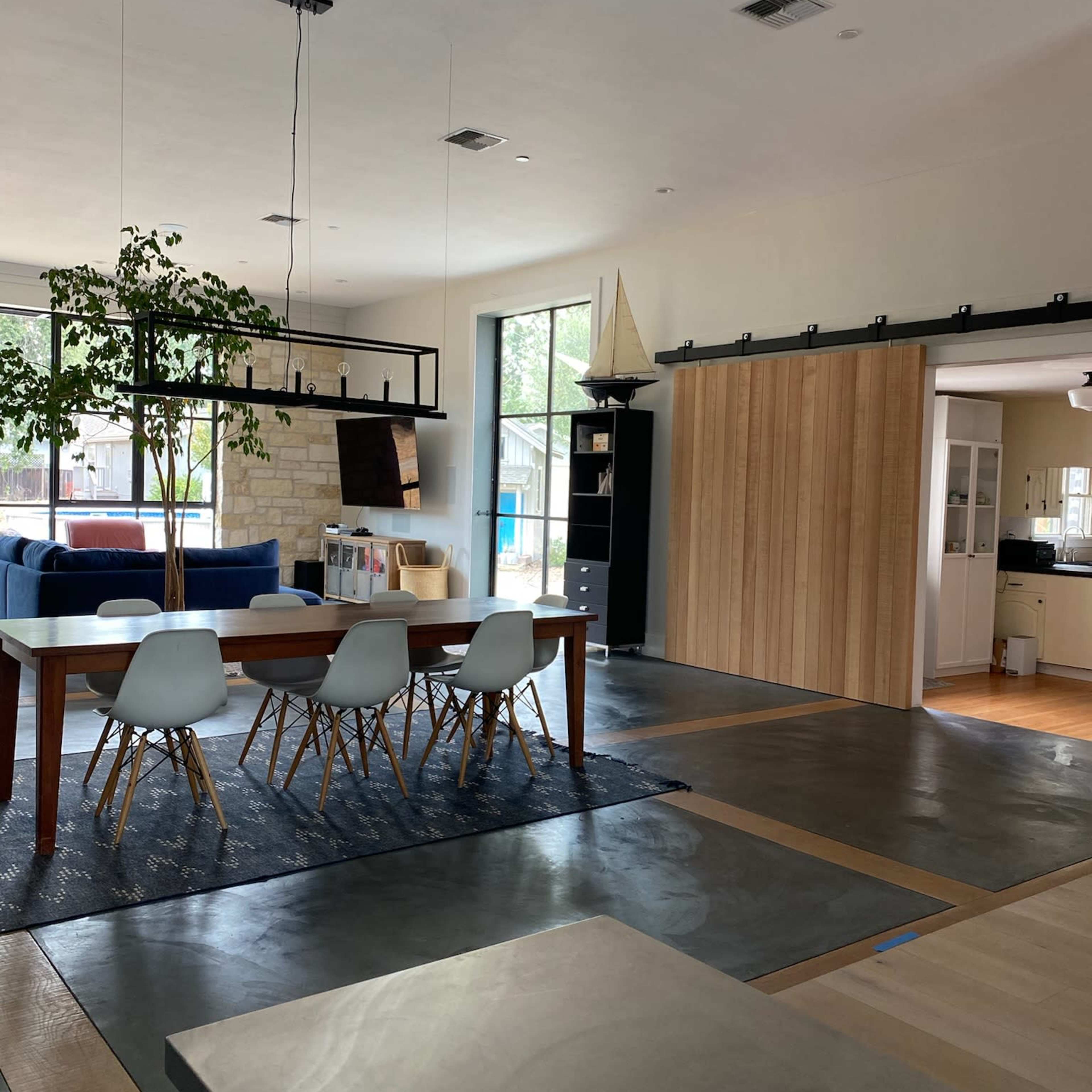 A modern interior space with a dining area featuring a wooden table and white chairs, a living area with a blue sofa, and a sliding barn door leading to a kitchen.