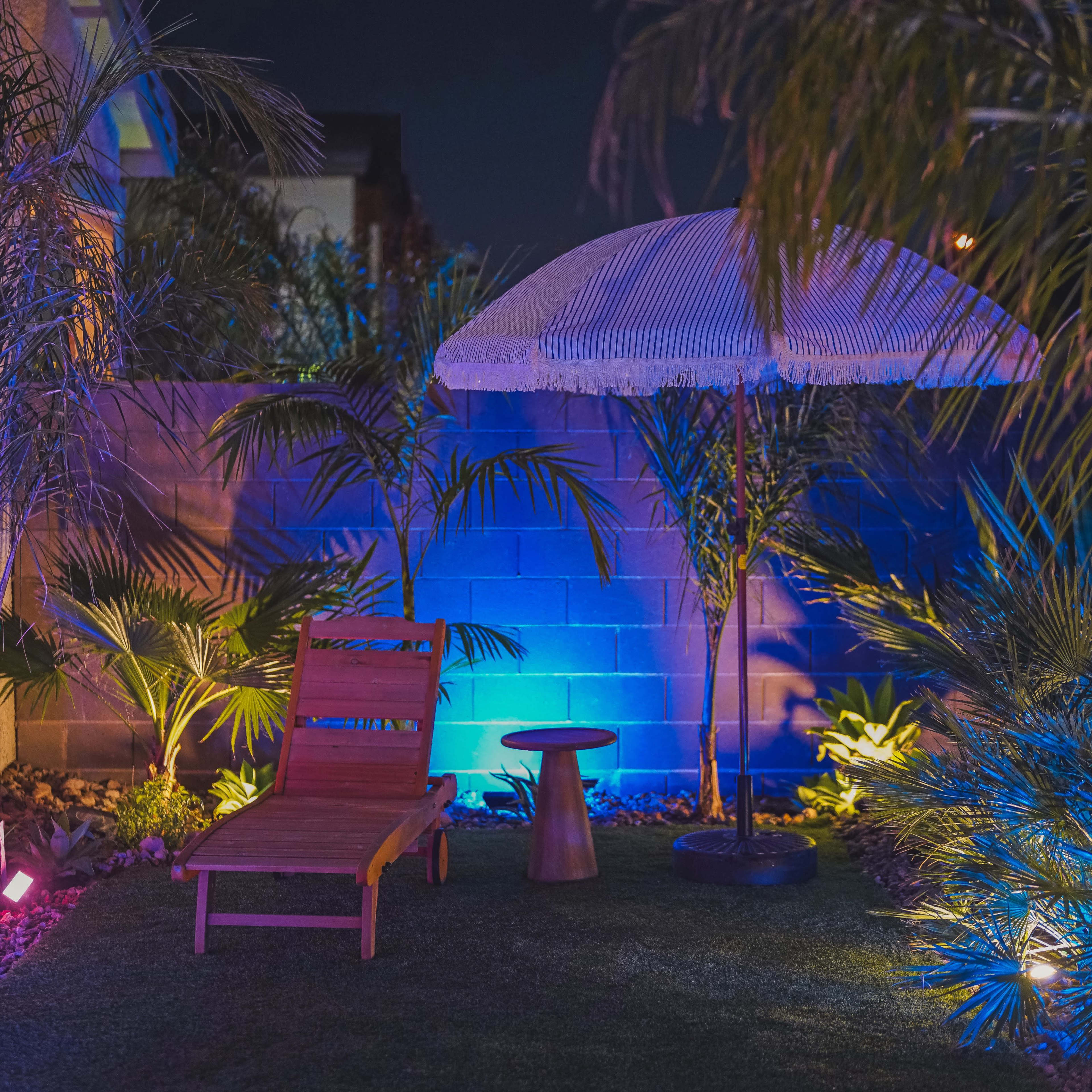 A wooden lounge chair and a small table sit beneath a large umbrella in a landscaped backyard illuminated by blue and purple lights.