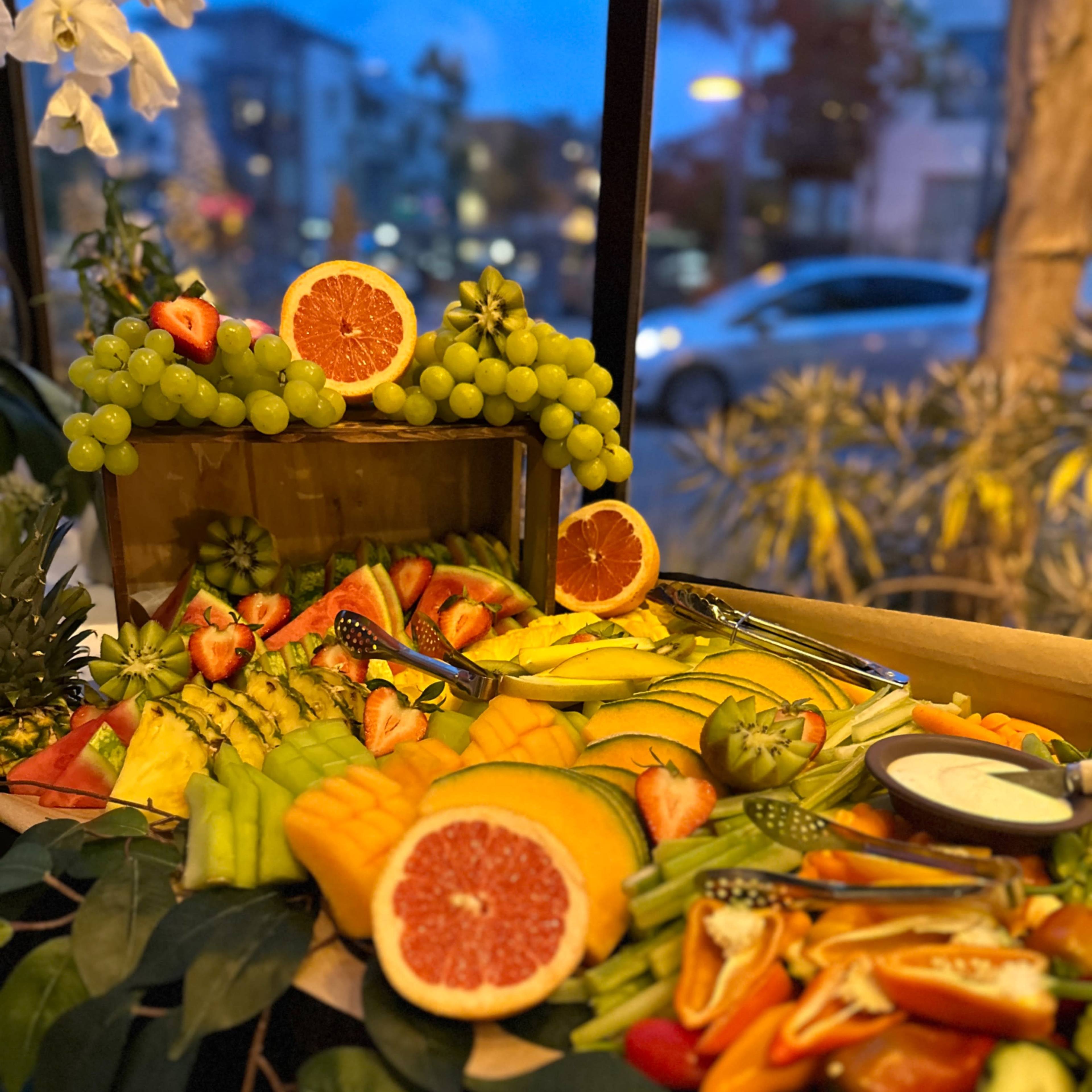 A colorful fruit platter featuring a variety of sliced fruits, including oranges, strawberries, grapes, and melons, arranged with greenery in a brightly lit setting.