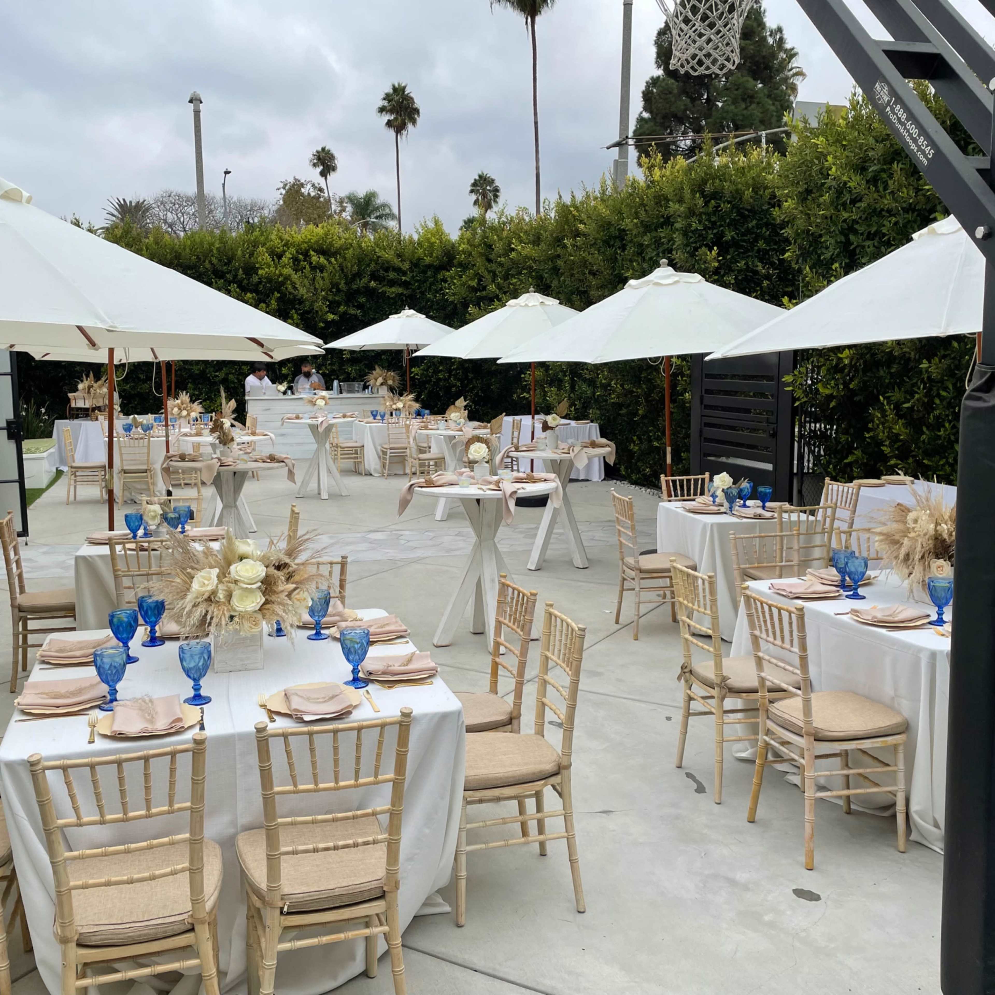 The outdoor dining area features several tables set with white tablecloths, blue glassware, and decorative elements, all under white umbrellas surrounded by greenery.