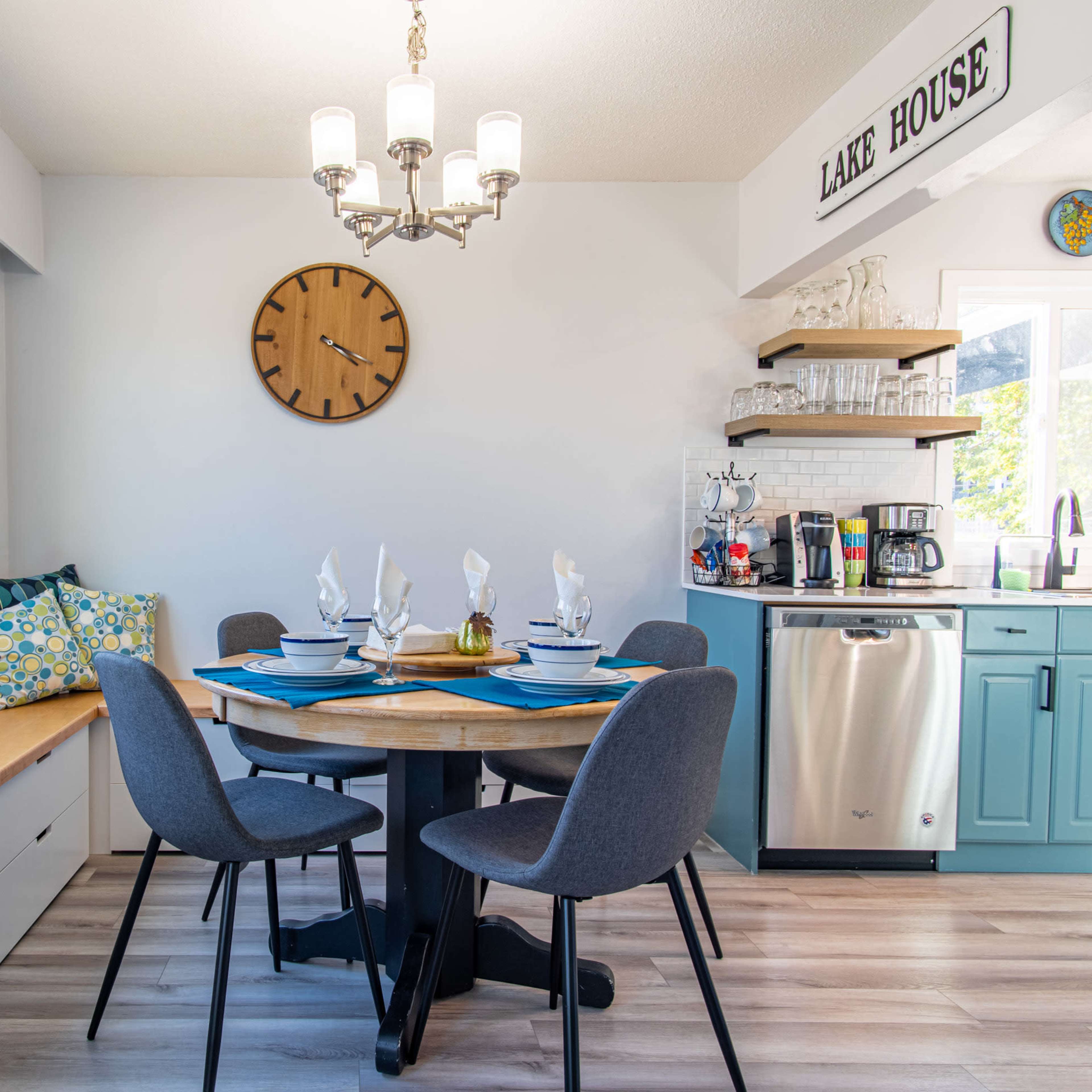 A dining area features a round table set for five, a built-in bench with cushions, and a wall clock above a sideboard in a room with large windows.