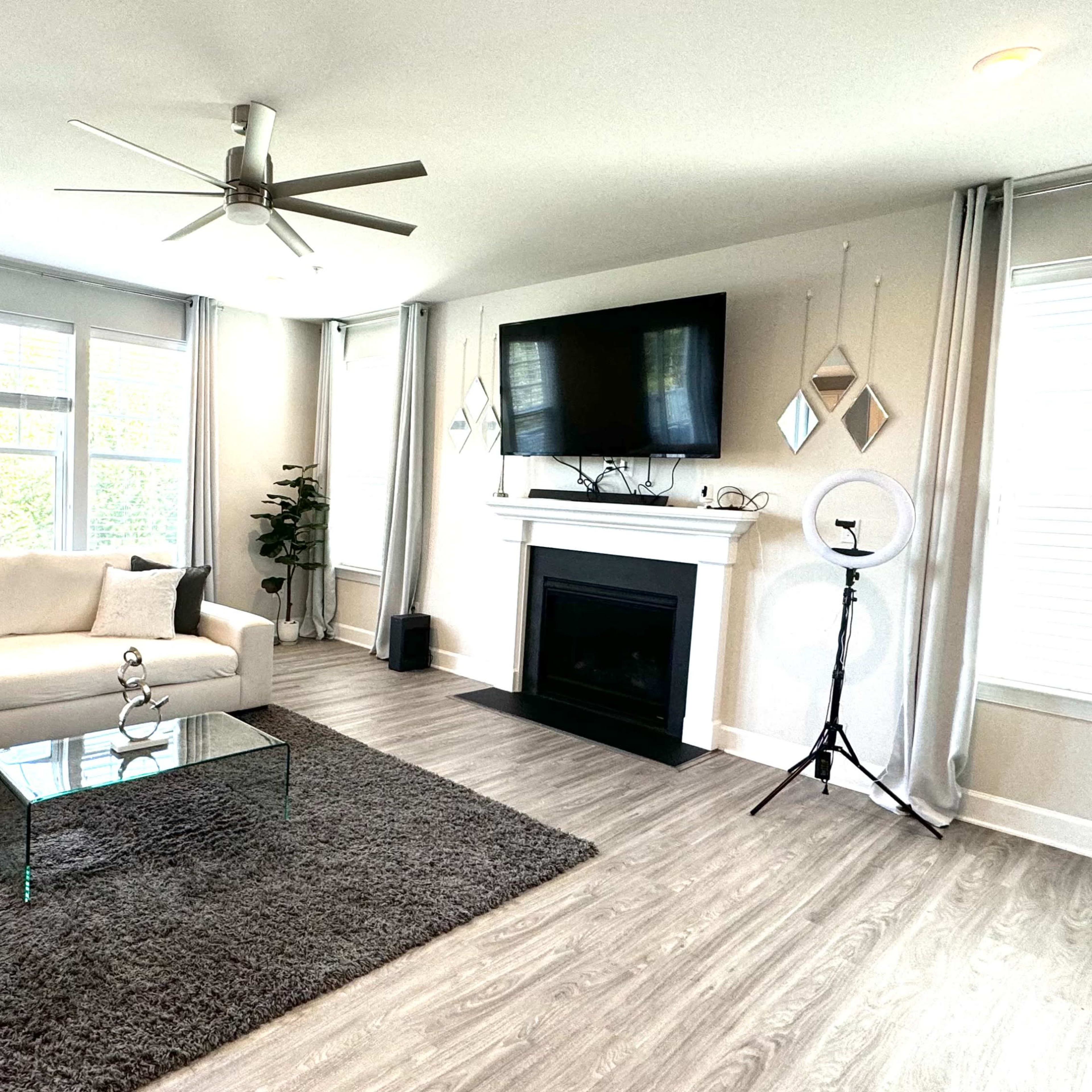 The living room features a white sectional sofa, a glass coffee table, a fireplace with a television above, and large windows with light curtains.