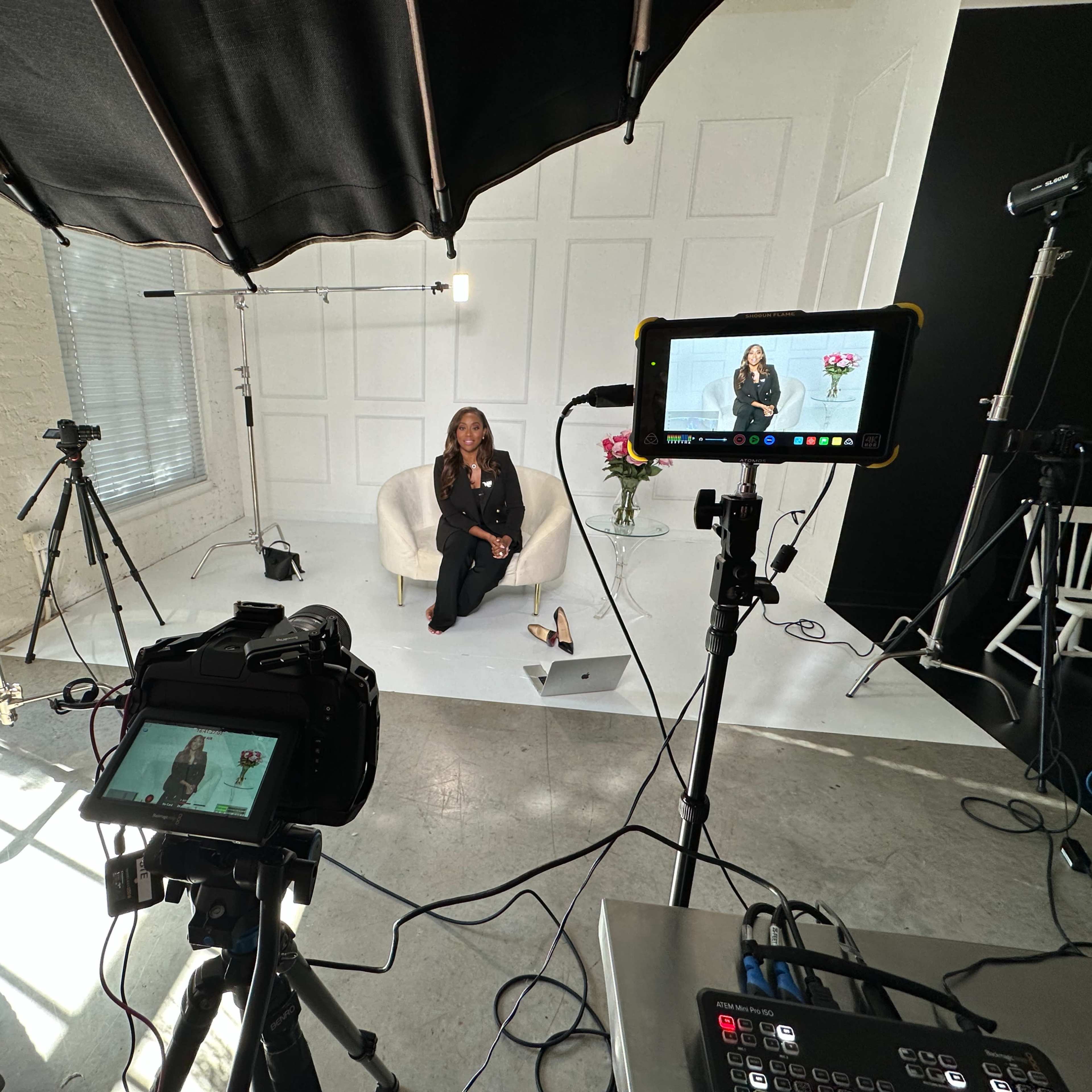 A woman sits on a white chair in a well-lit studio surrounded by multiple cameras and lighting equipment.