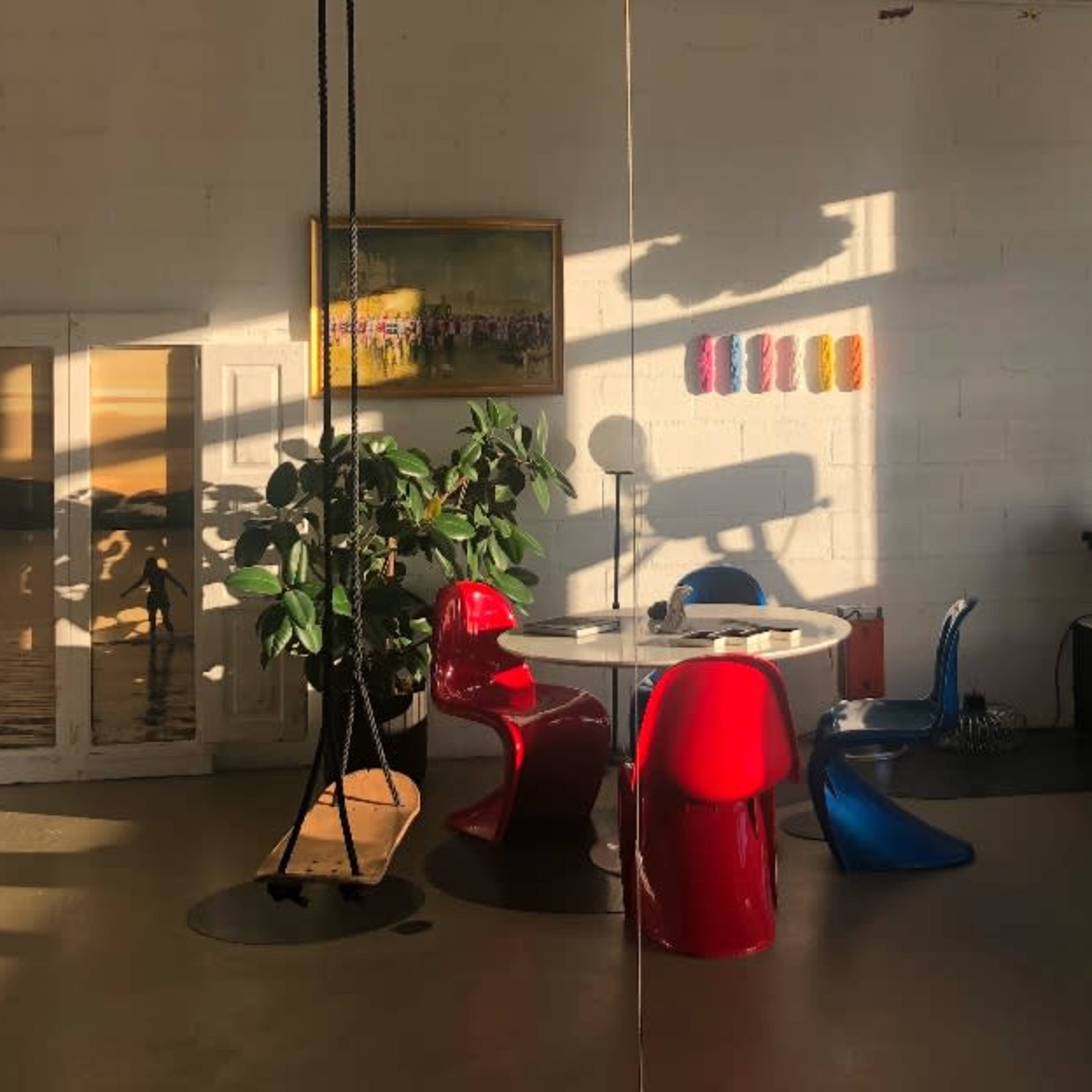 A modern, brightly lit room with a round table surrounded by colorful chairs, a potted plant, a mannequin, and various decorative elements along the window.