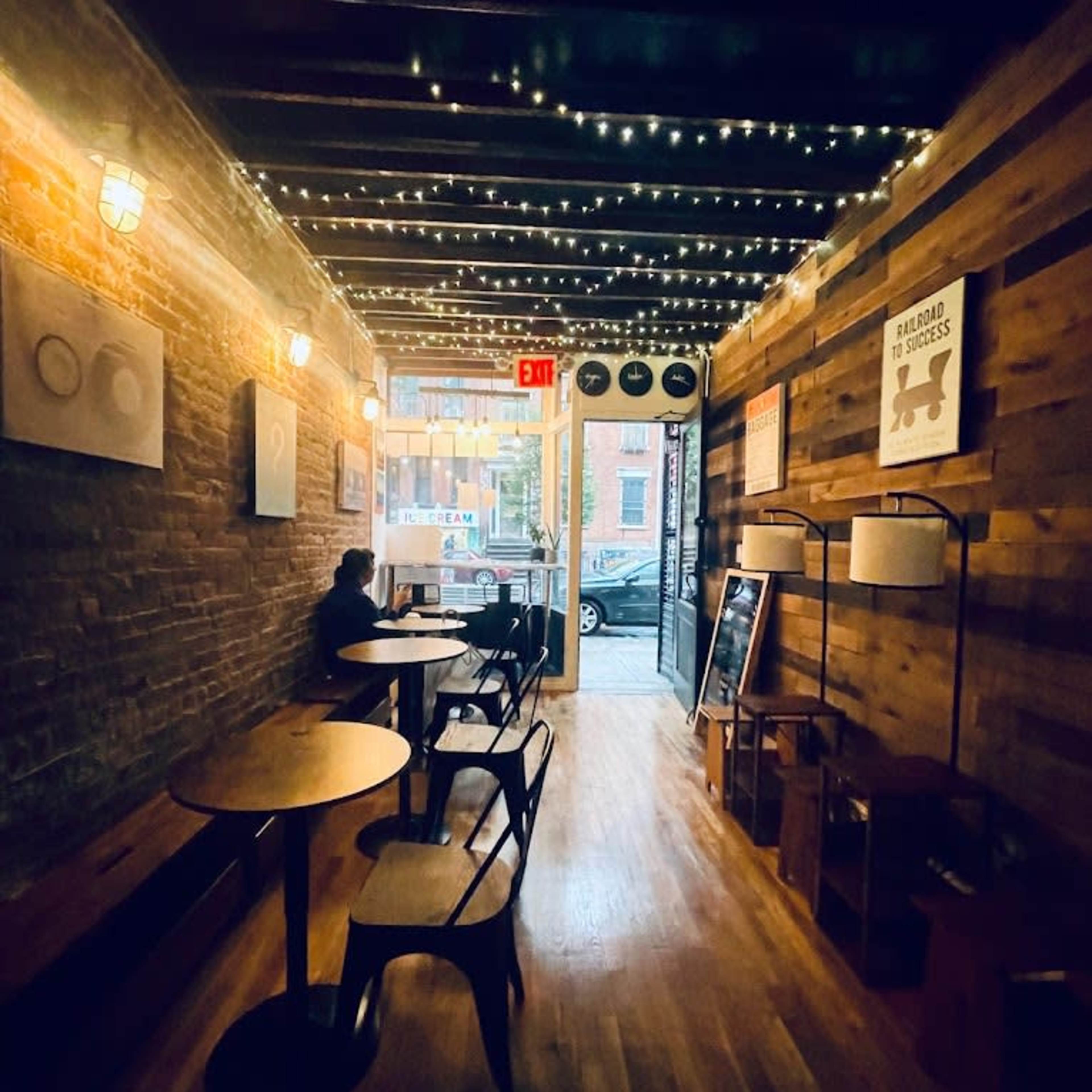 A dimly lit hallway of a café with wooden walls, small tables, and string lights overhead, leading to an exit that opens to the street.