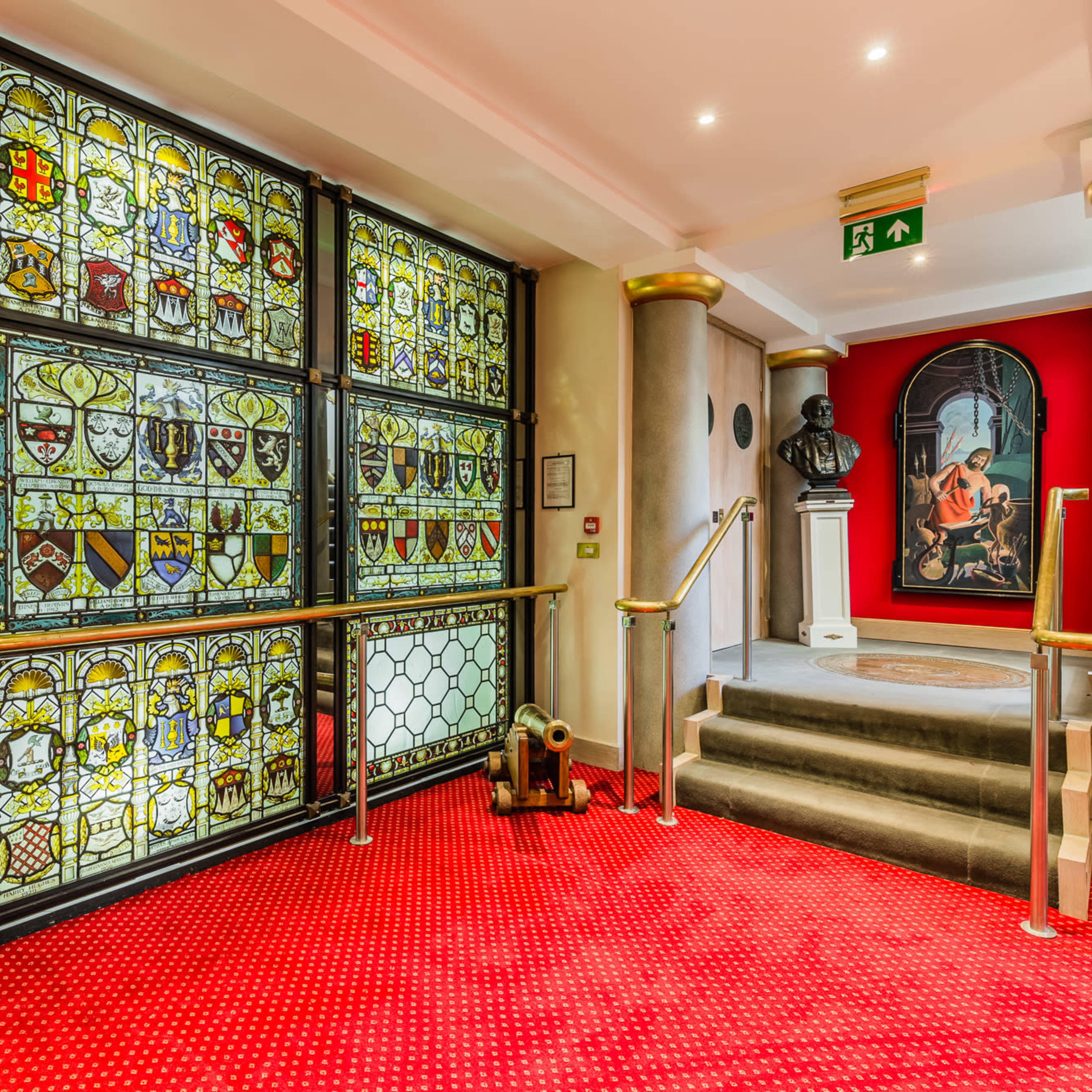 The scene features a hallway with ornate stained glass windows displaying various heraldic shields, leading to a staircase beside a decorative bust and a framed painting on a red wall.