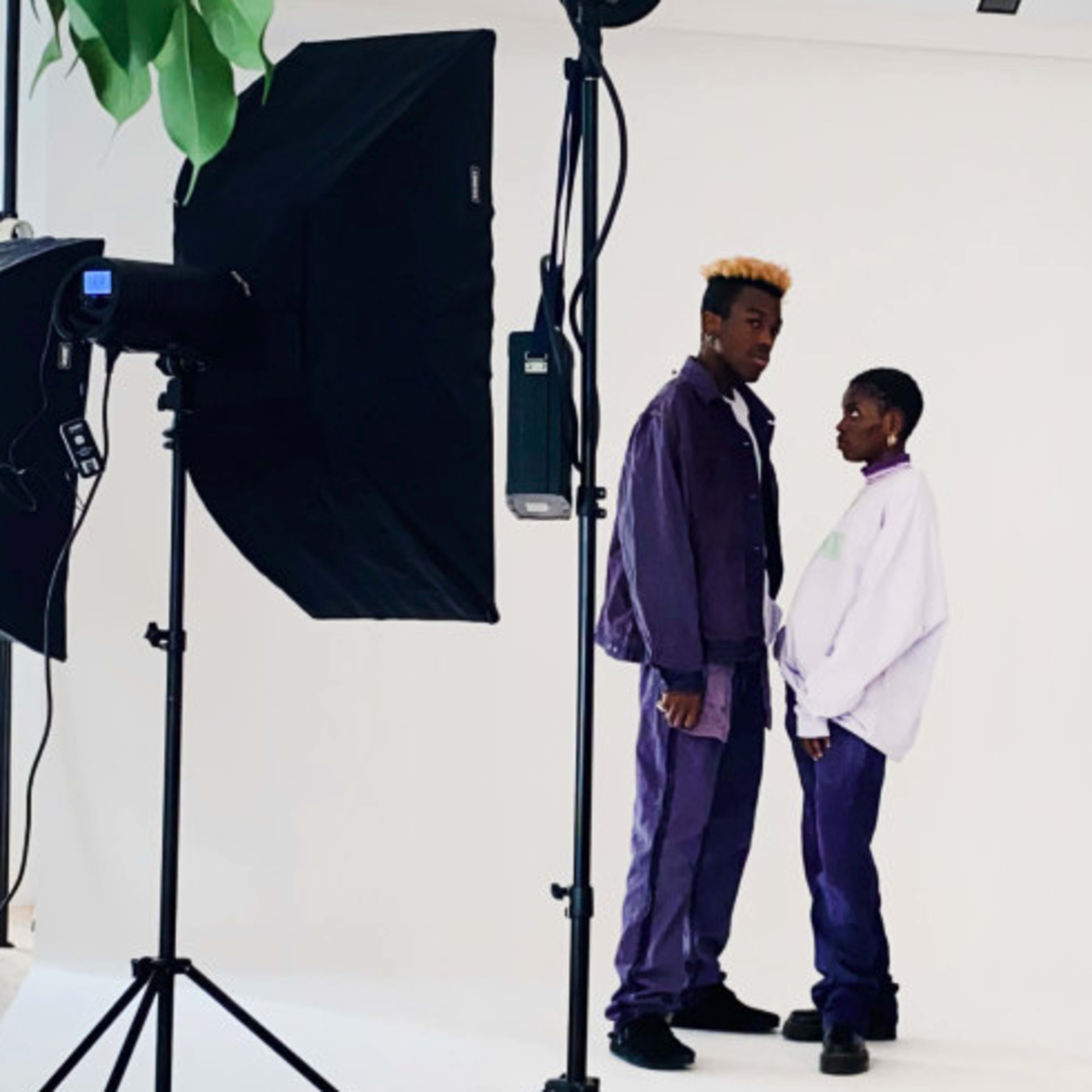 Two individuals pose for a photo in a studio setting, surrounded by professional lighting equipment and a plain background.