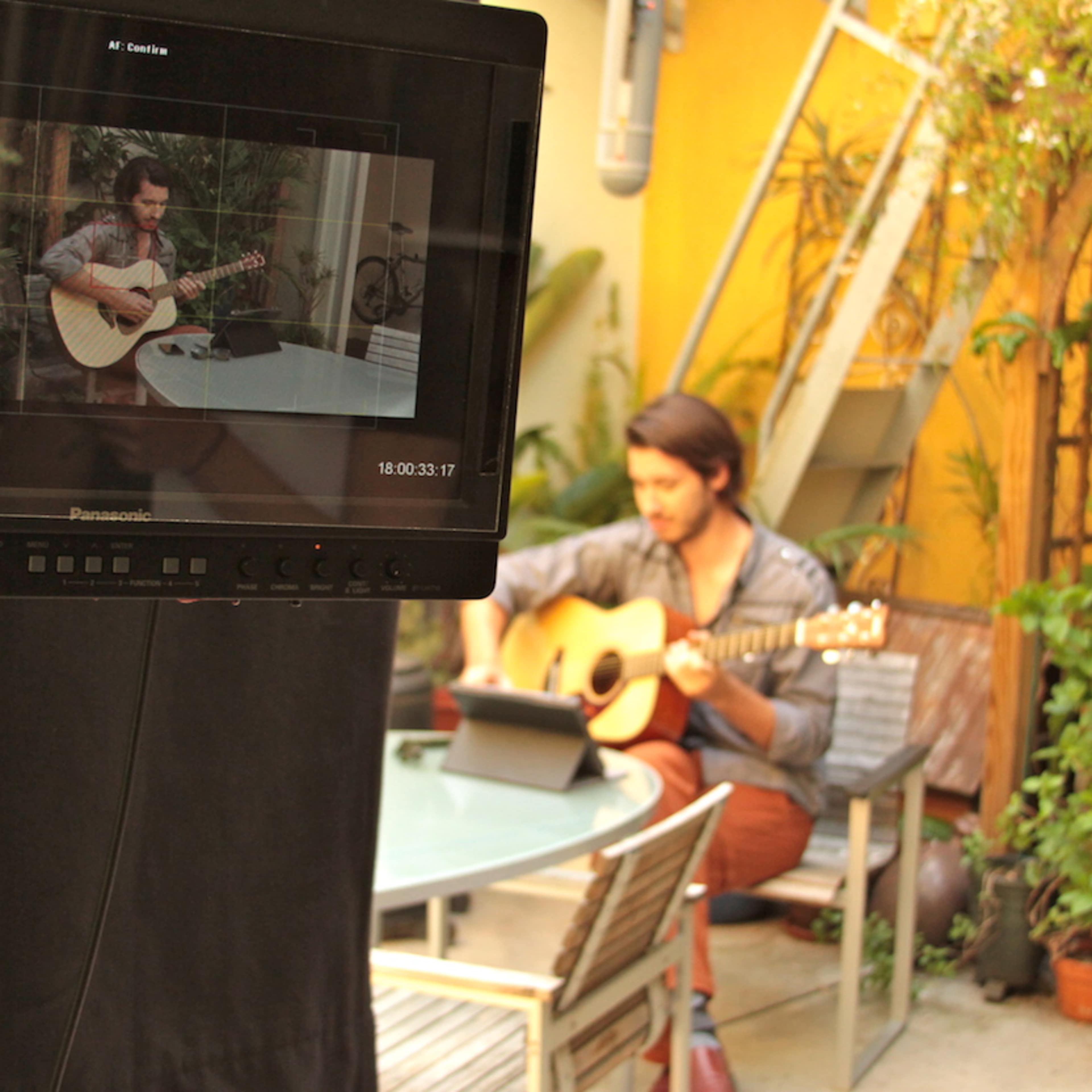 A man is playing a guitar in a lush outdoor setting, with a camera monitor displaying the scene in front of him.