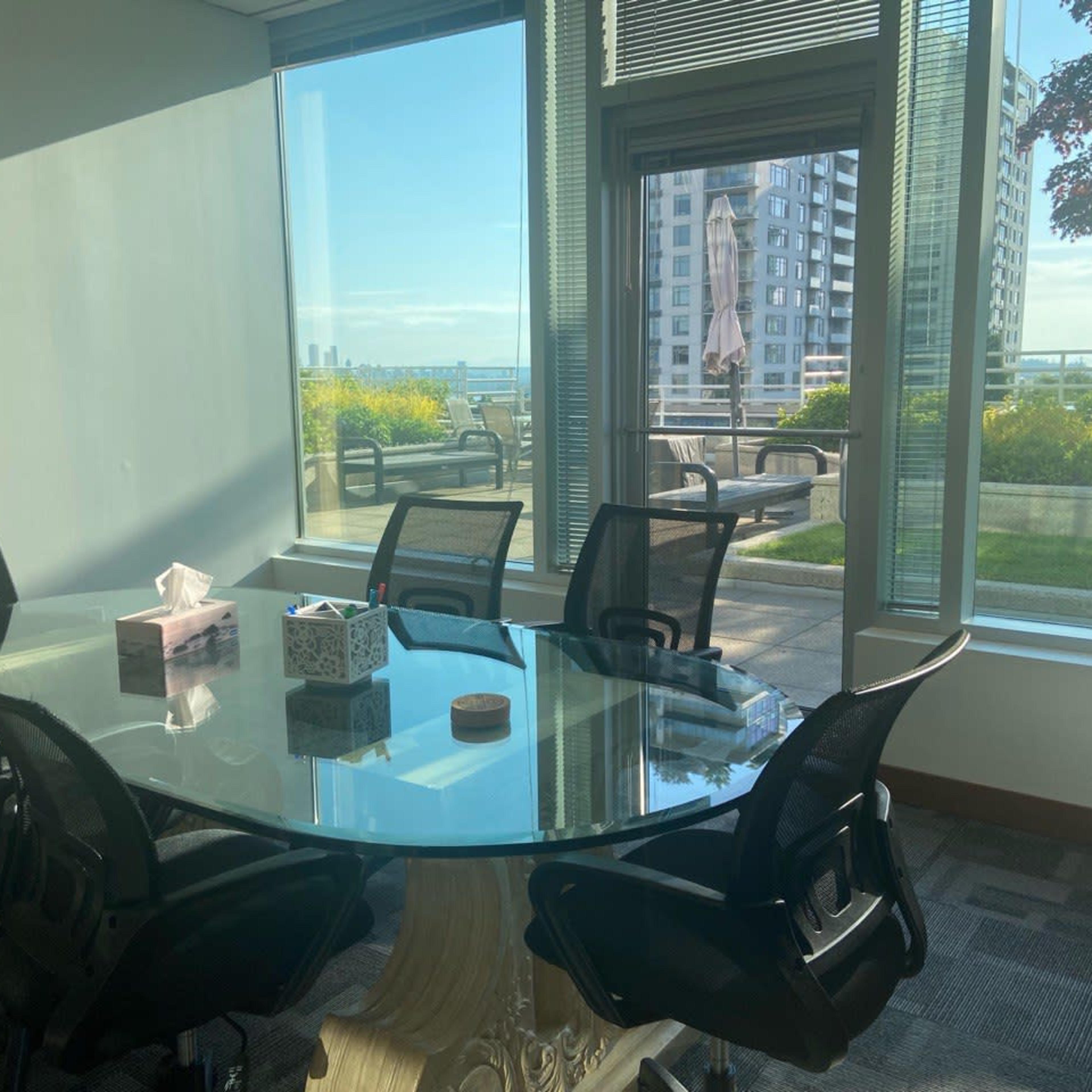 A modern conference room features a glass table surrounded by black chairs and large windows overlooking a green outdoor terrace and city skyline.