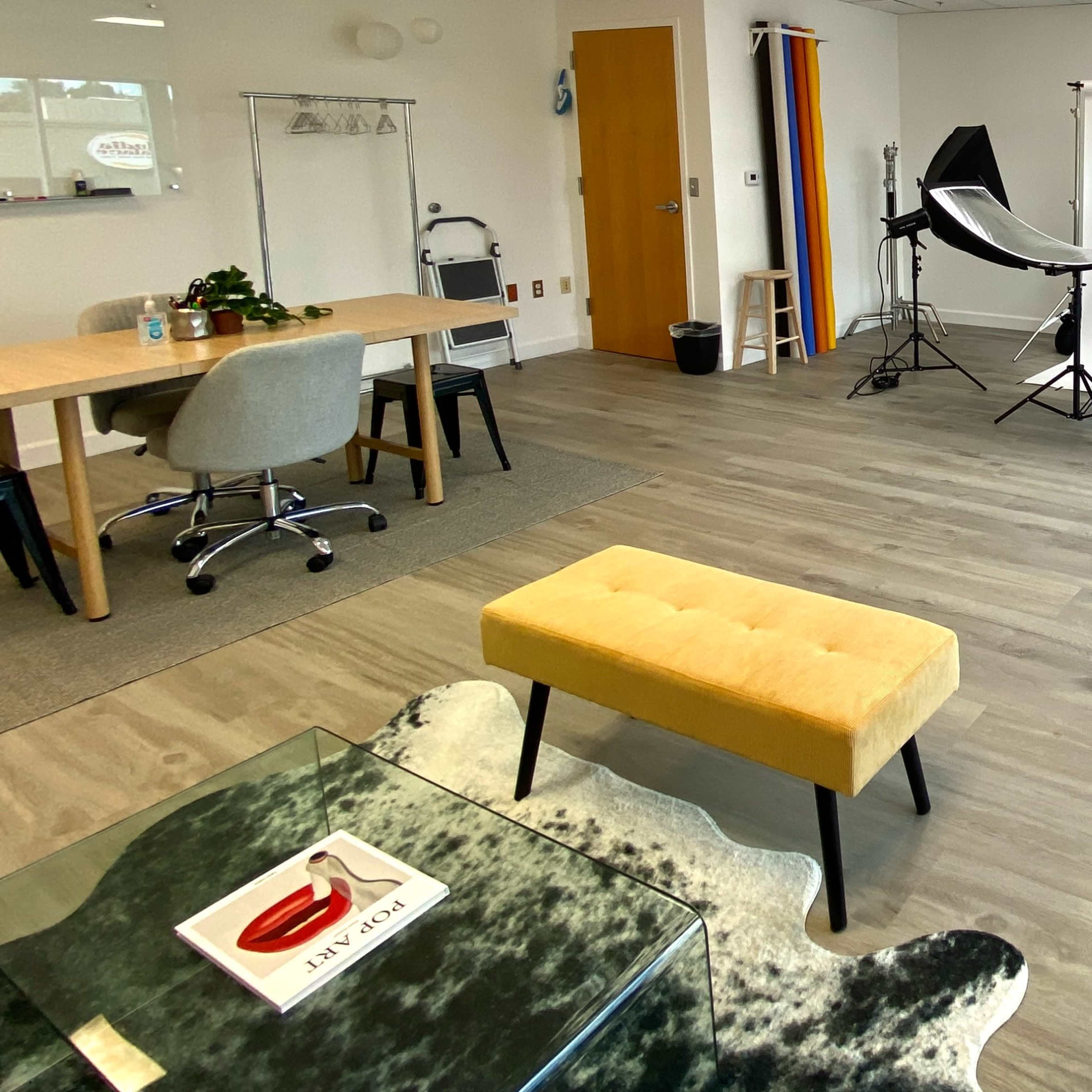 A well-lit studio space set up for photography, featuring backdrop equipment, lighting, and a seating area with a yellow bench and a glass table.