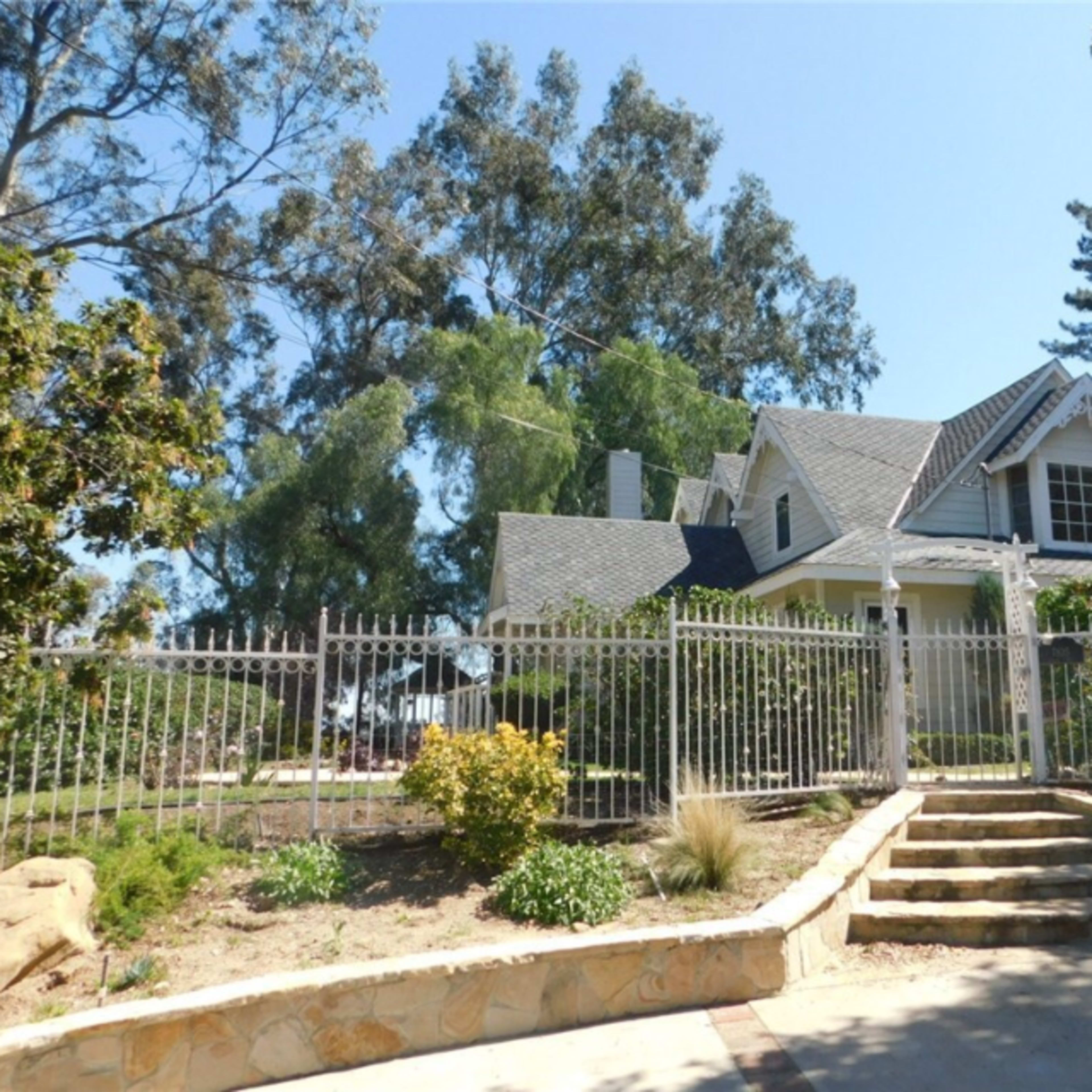 A large, two-story house is surrounded by a wrought iron fence and set among tall trees and landscaped gardens.