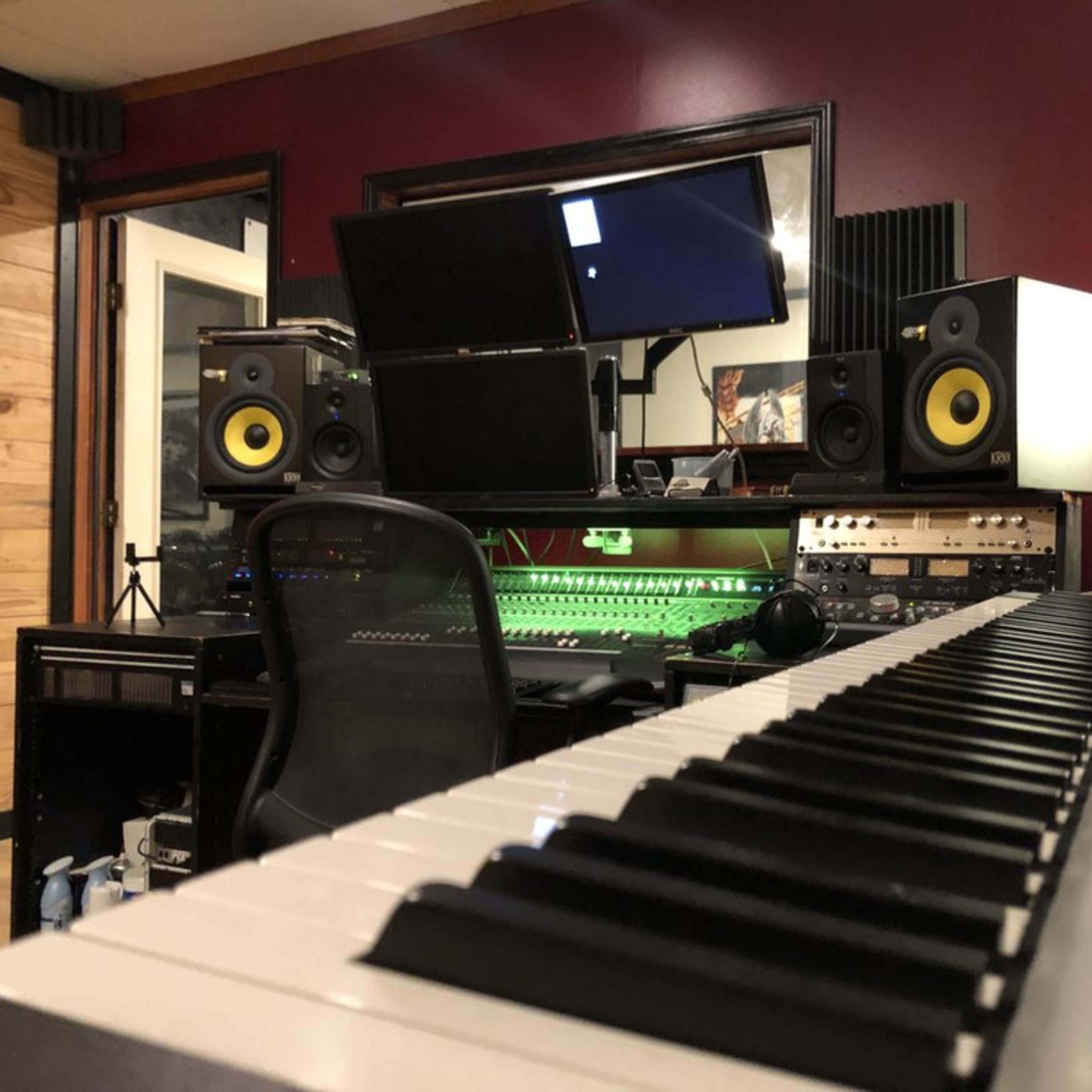 A music studio with a keyboard in the foreground, a mixing console, dual computer monitors, and studio speakers in the background against a red wall.