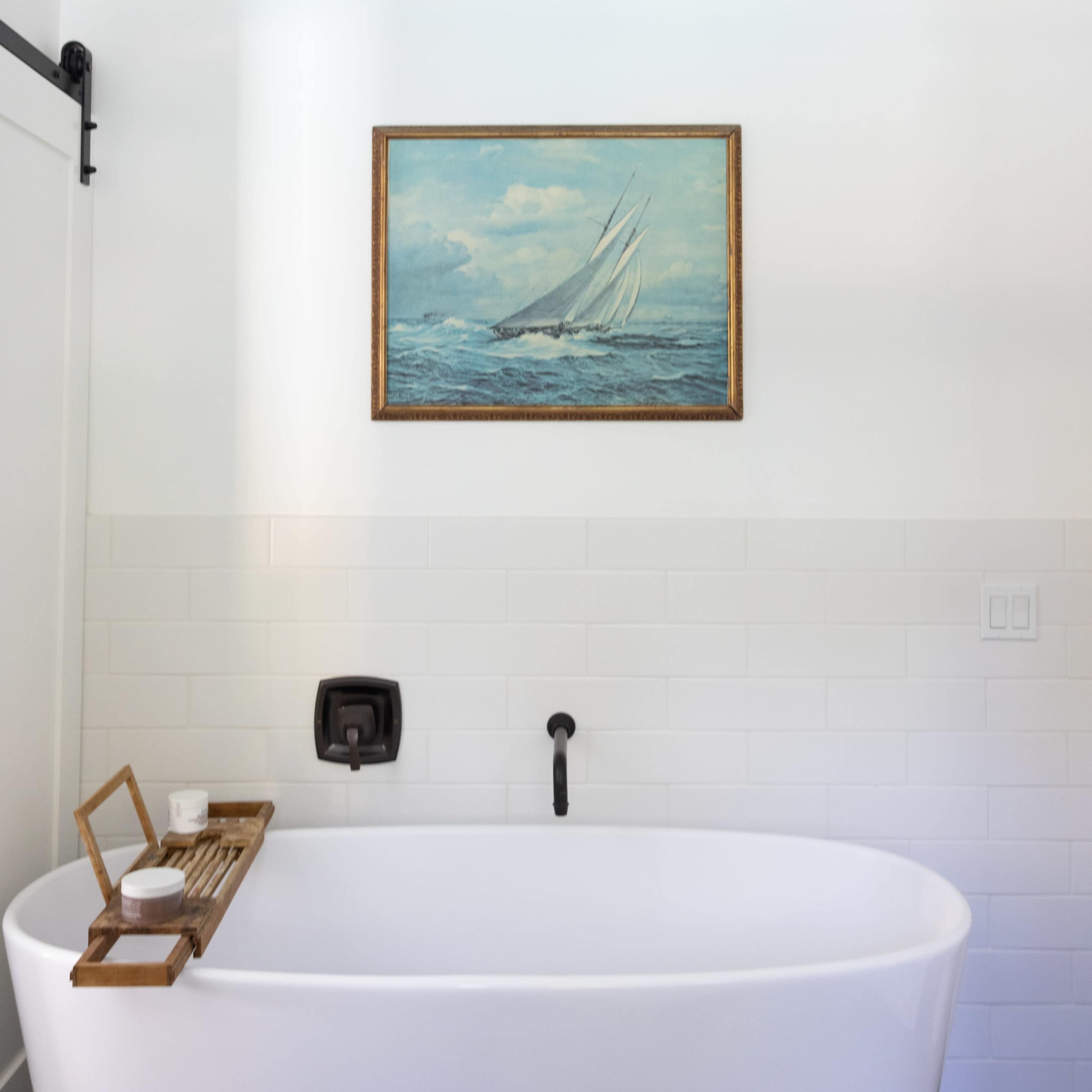 A modern bathroom featuring a freestanding bathtub, a wooden bath caddy, and a landscape painting of a sailing ship mounted on the wall.