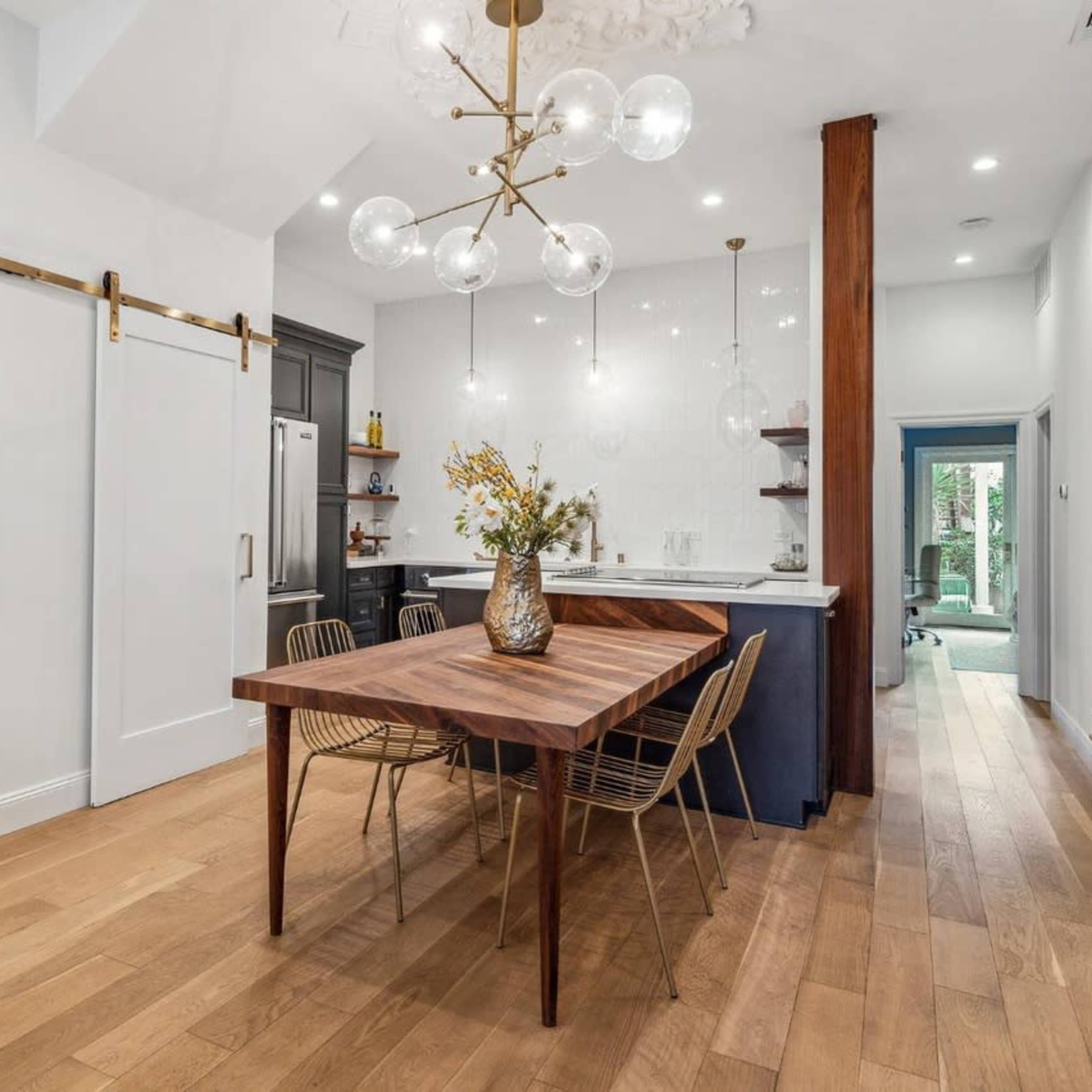 A modern dining area featuring a wooden table with metal chairs, a large light fixture overhead, and a kitchenette in the background.