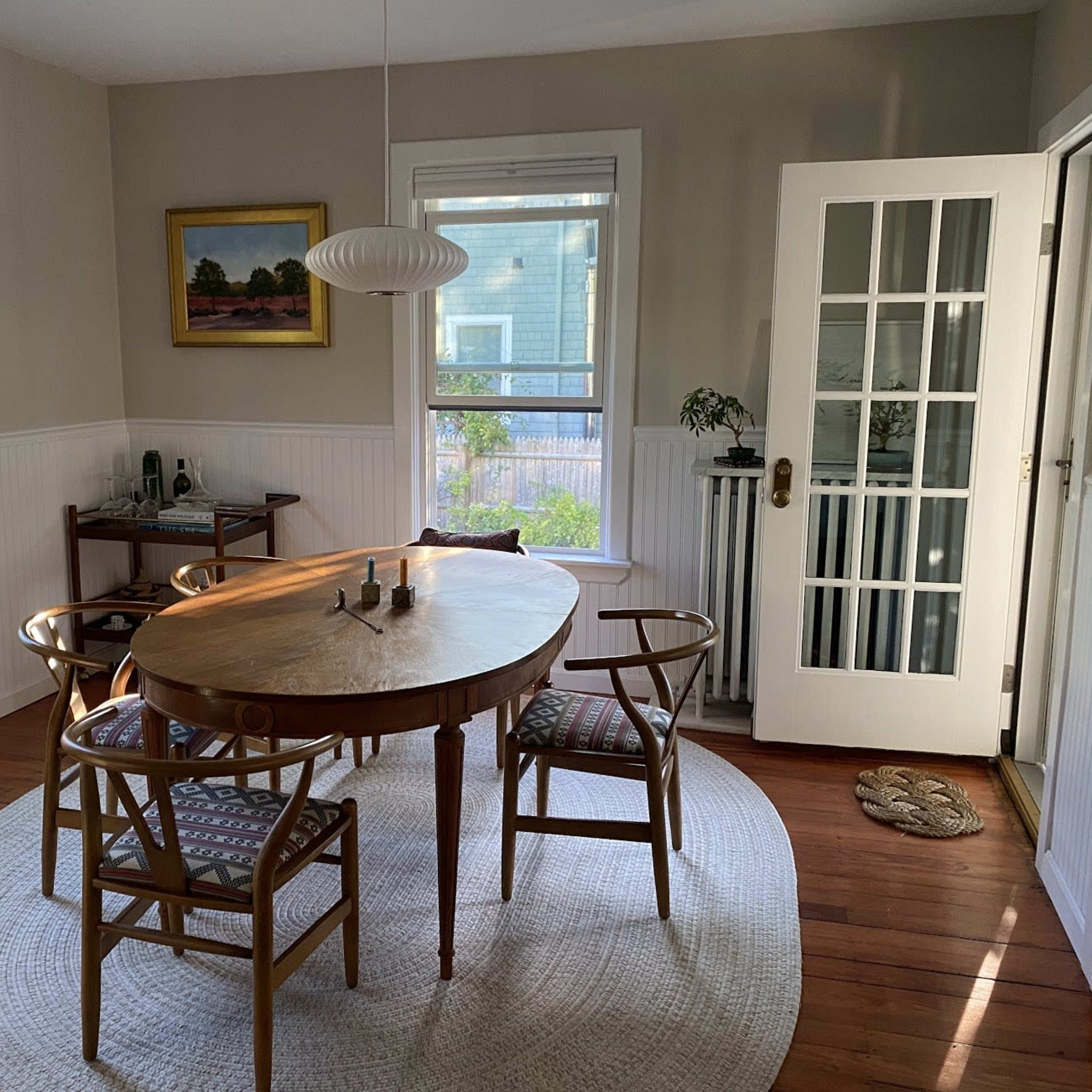 A round wooden dining table surrounded by four chairs is set in a light, airy dining room with large windows and a door leading outside.
