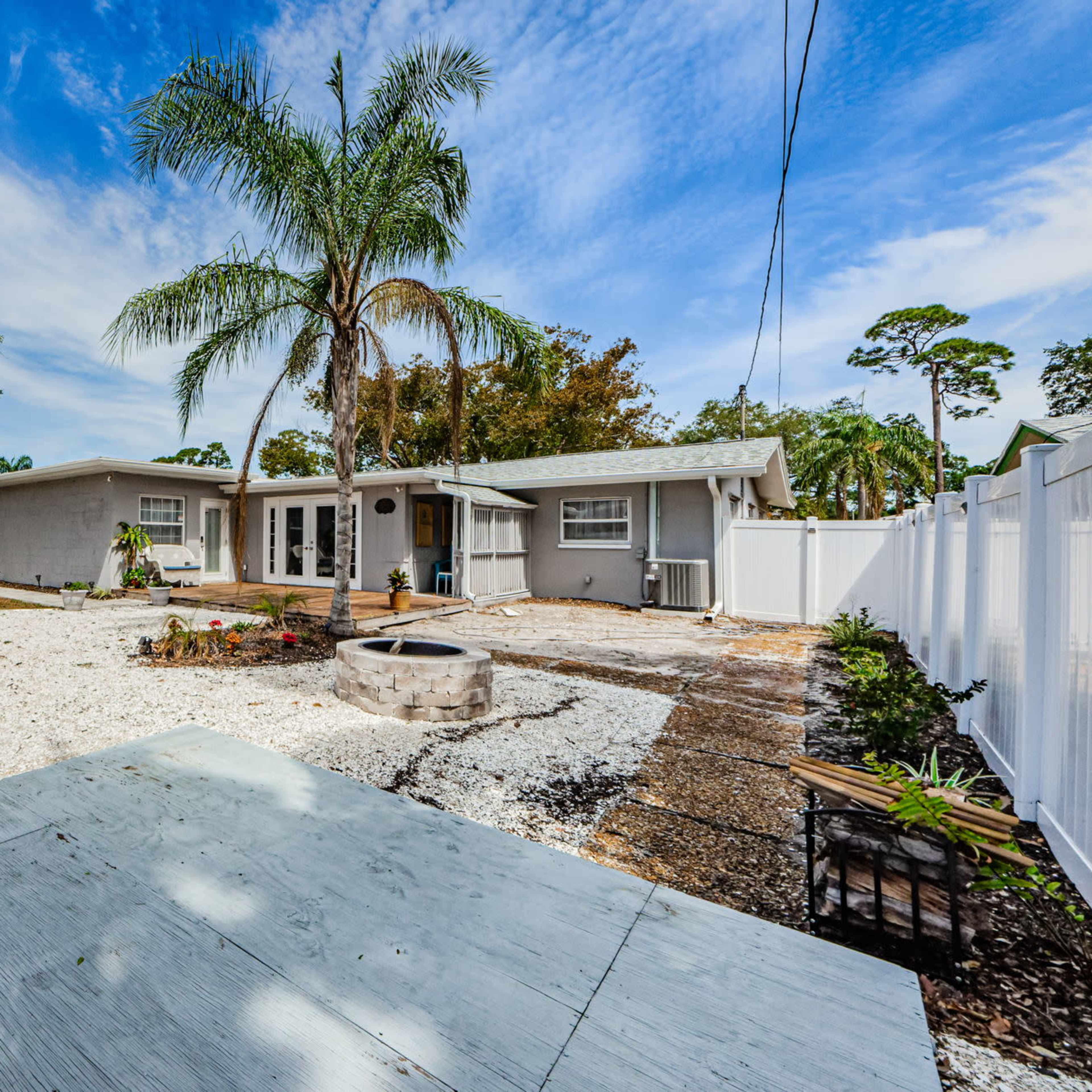 A spacious backyard with a gravel area, a circular fire pit, and a house surrounded by trees and a fence.