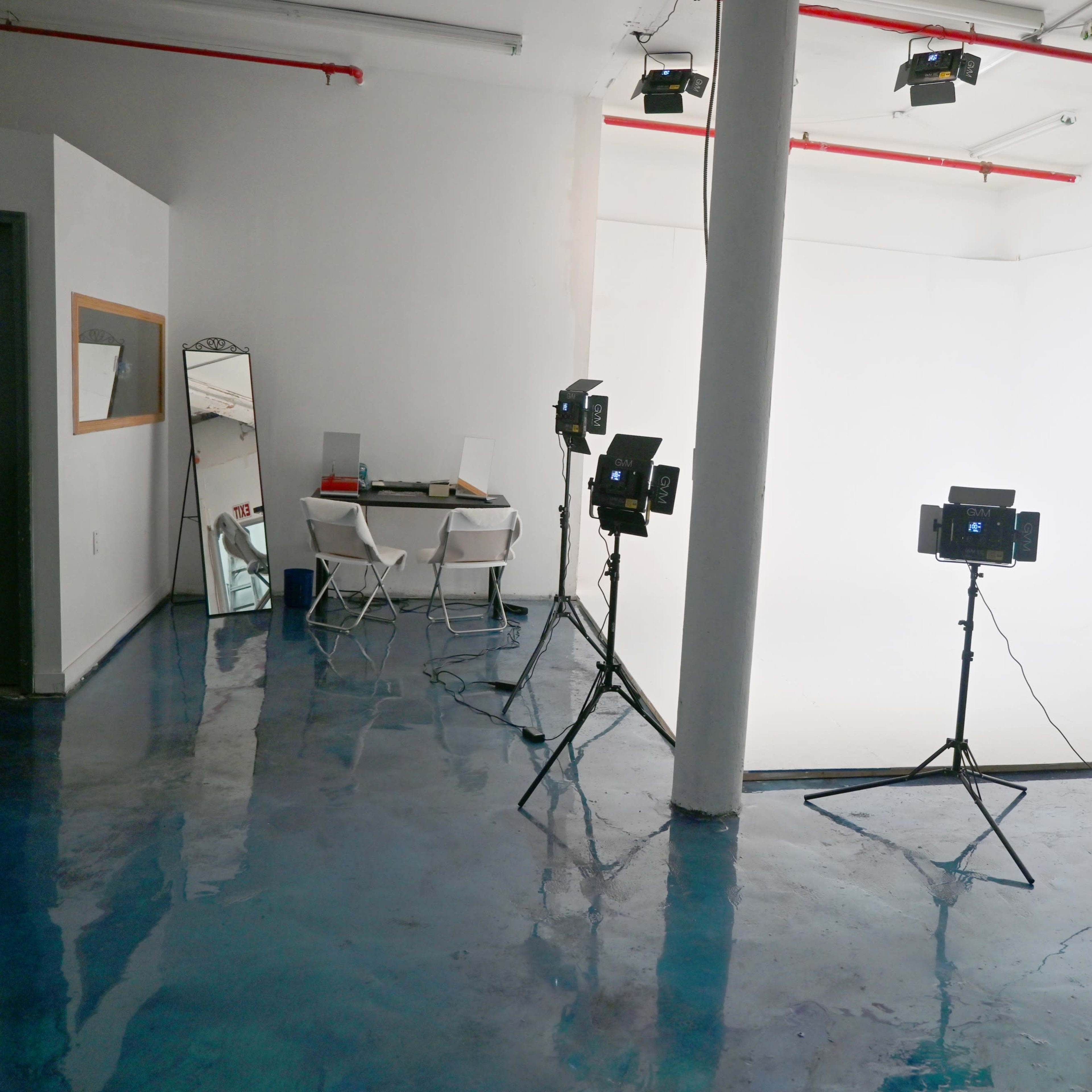 A photography studio with three light stands, a mirror, and a table with chairs against a white backdrop.