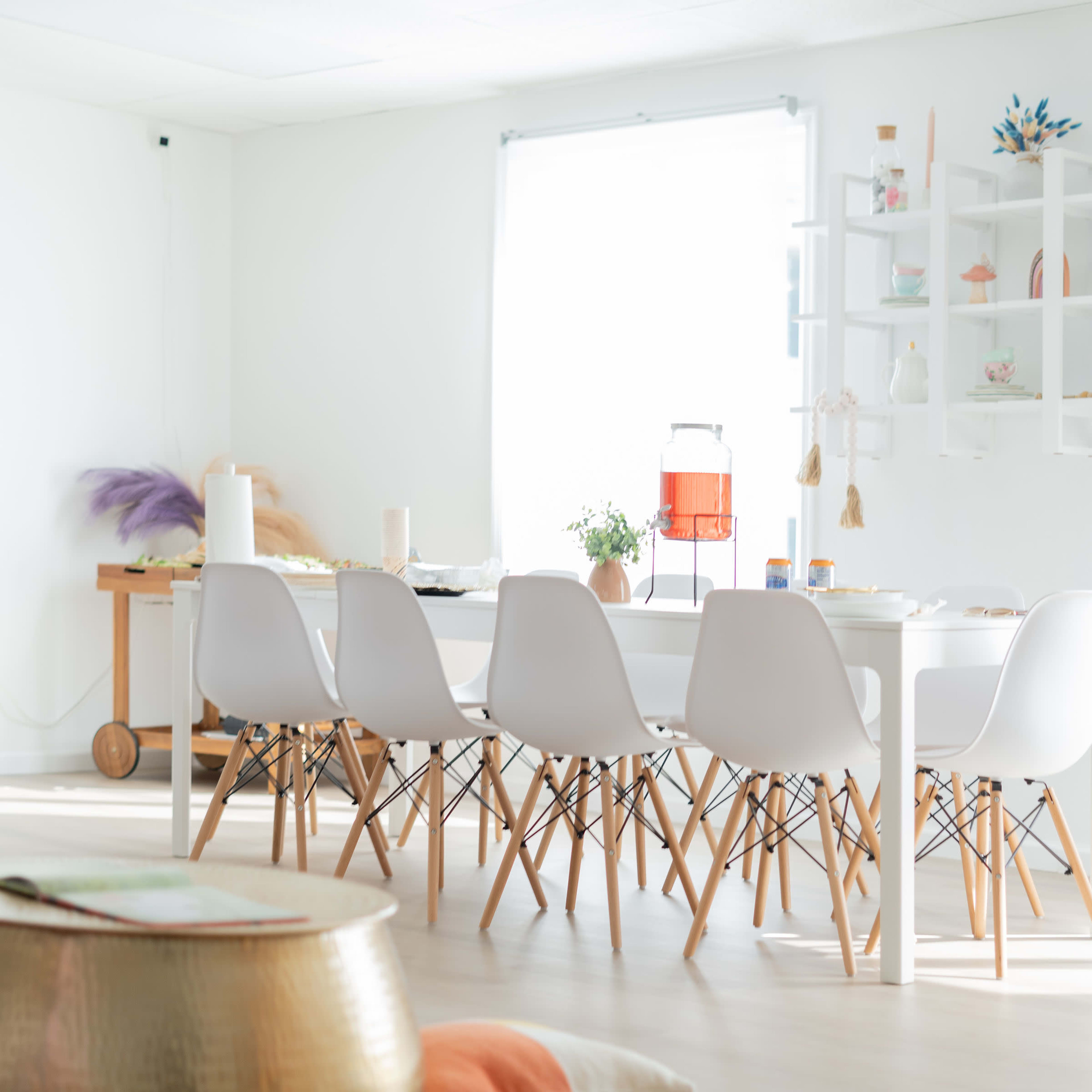 A bright, minimalist dining area features a large white table surrounded by chairs, a rug with colorful cushions, and decorative elements on the shelves.
