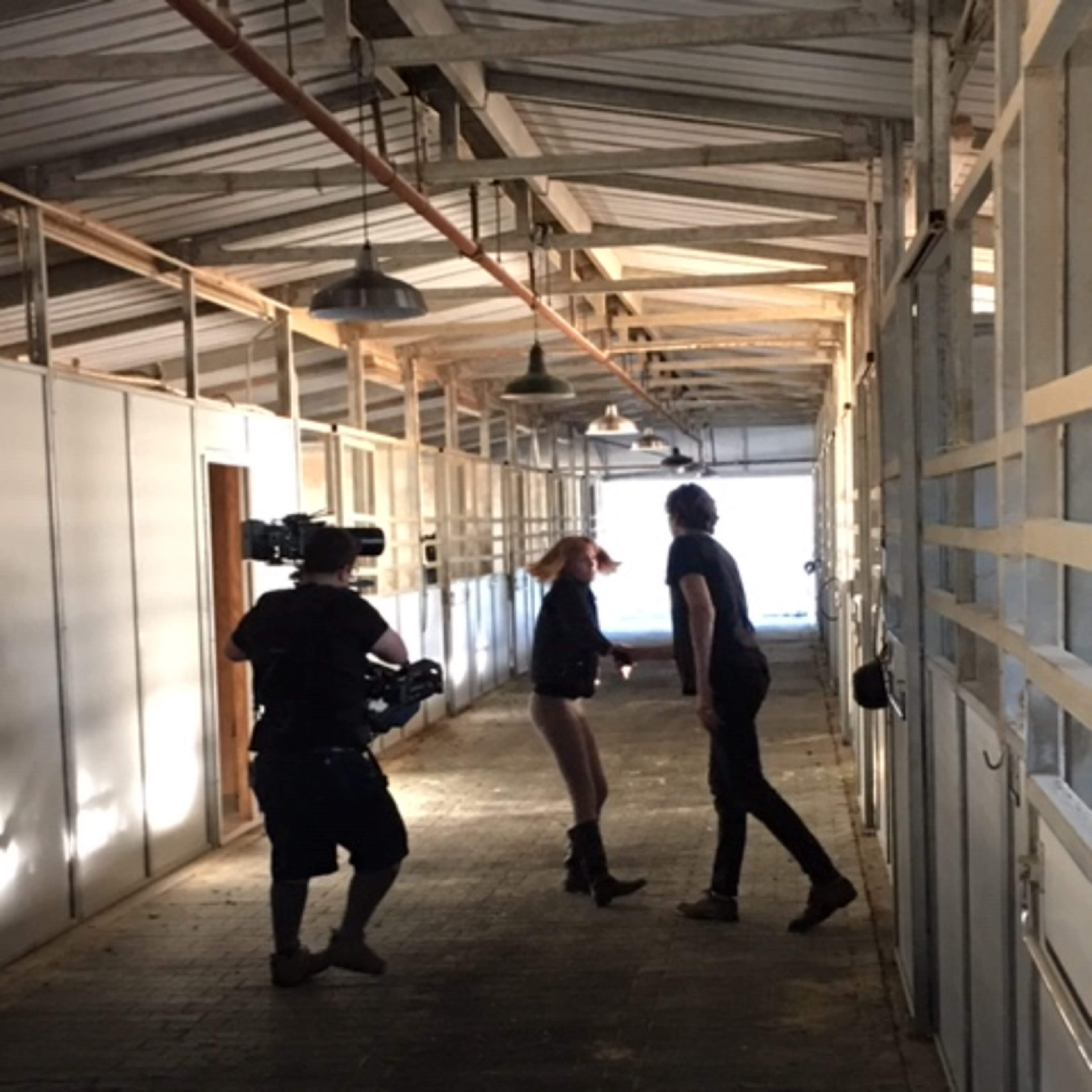 A camera operator records two people interacting in a well-lit corridor lined with horse stalls.