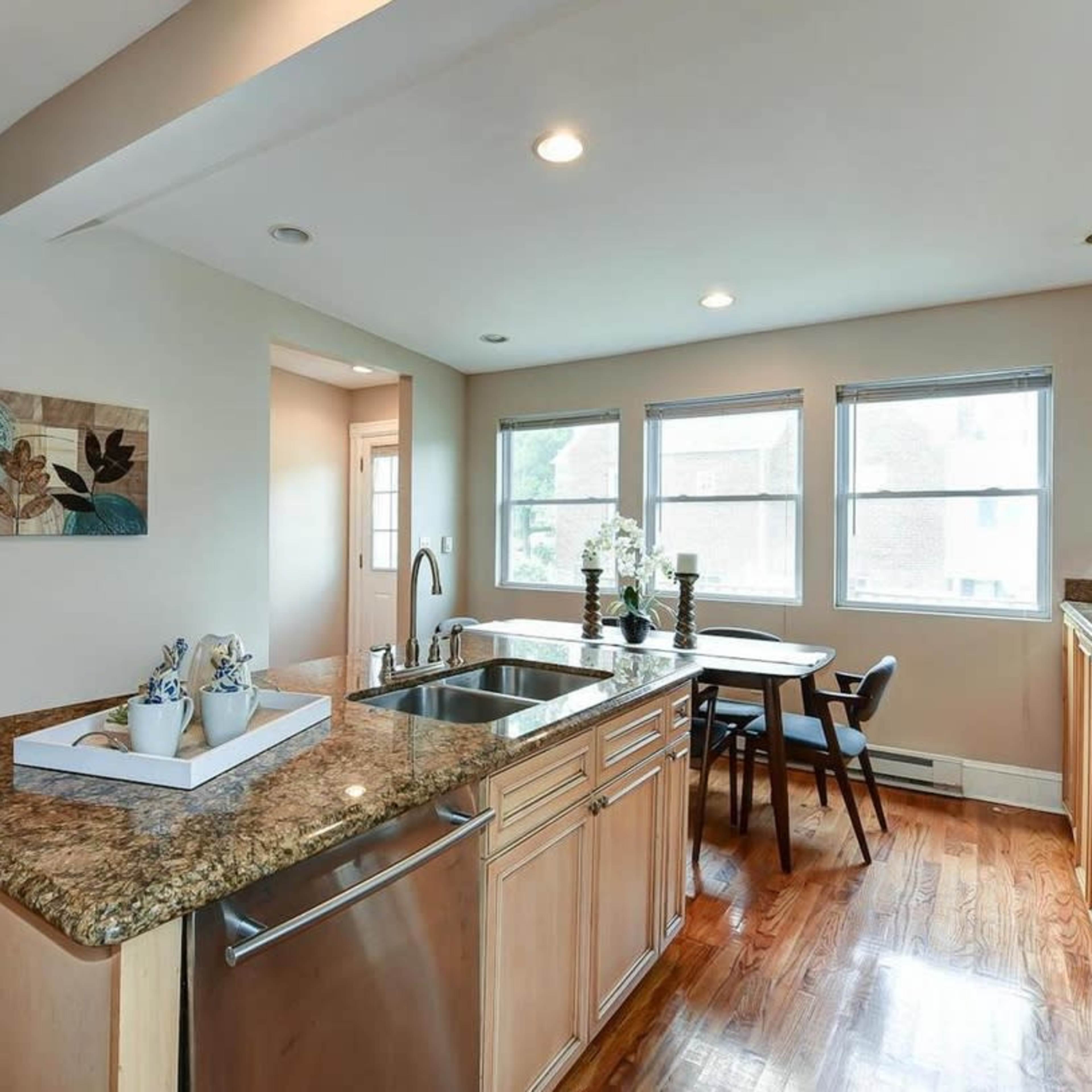 A modern kitchen features a granite countertop, sink, and a dining area with a table and chairs beside large windows.