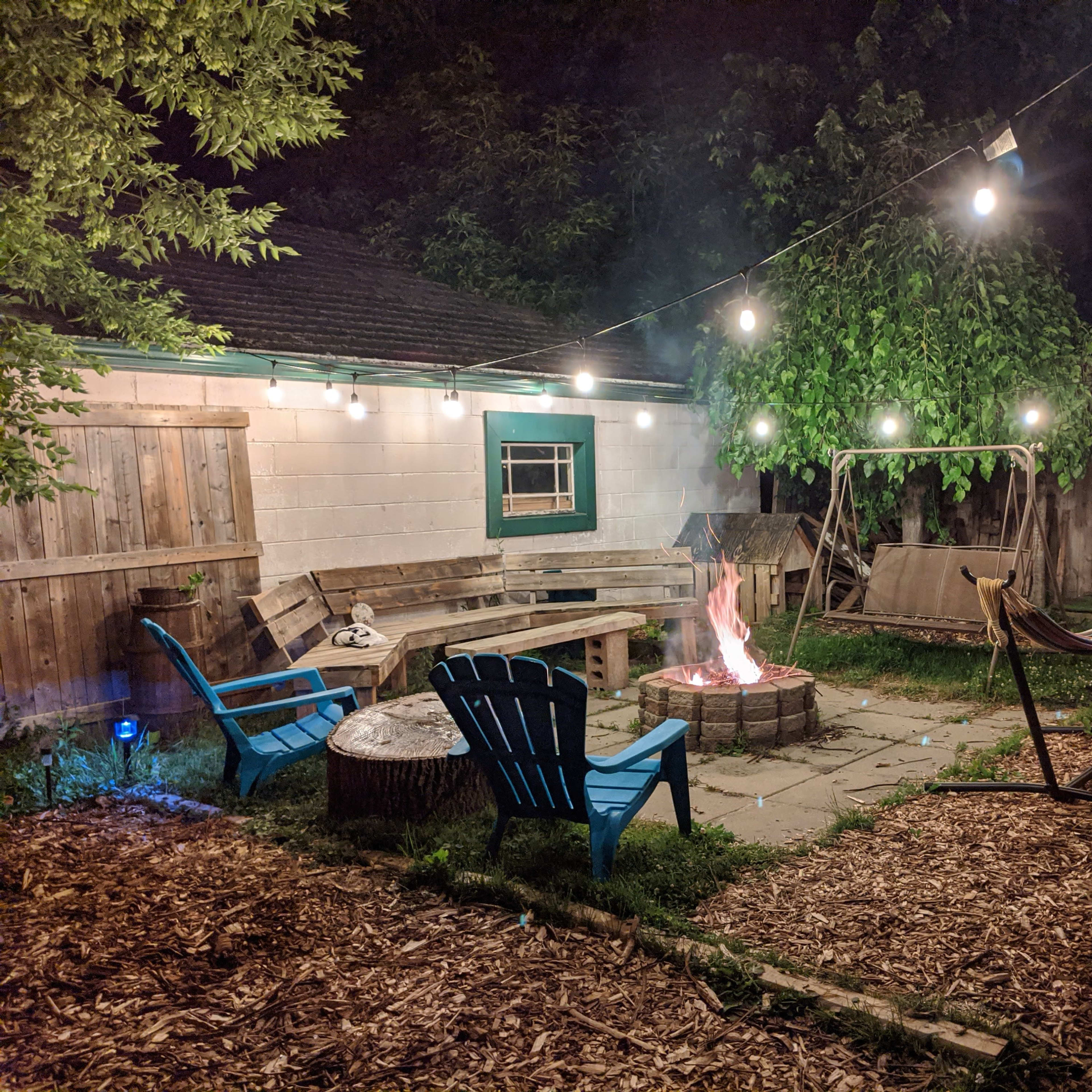 A backyard scene features a fire pit surrounded by seating, illuminated by string lights overhead, with a hammock and swing in the background.