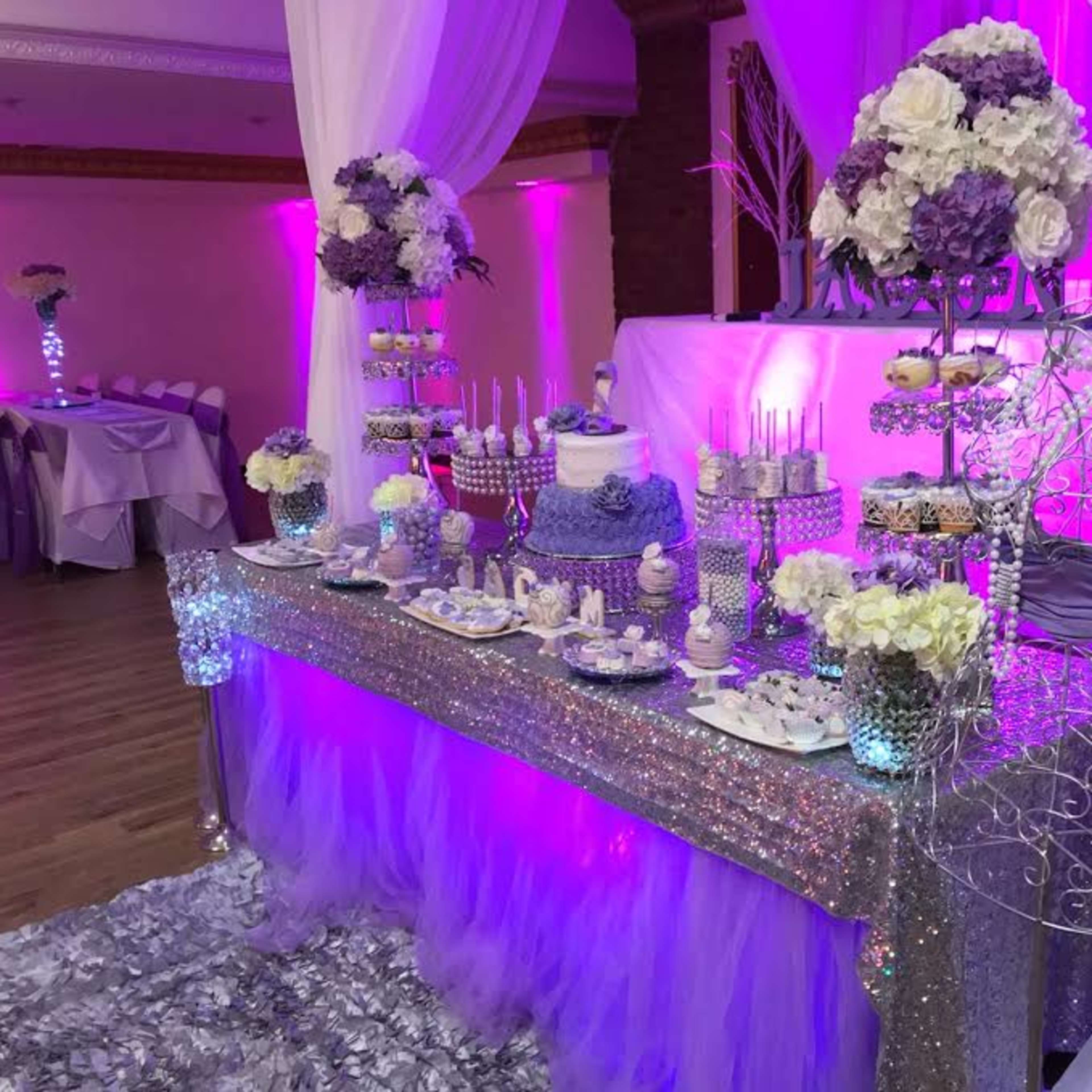 A decorated dessert table featuring a lavender cake, various sweets, and floral arrangements, set up in a venue with purple lighting.