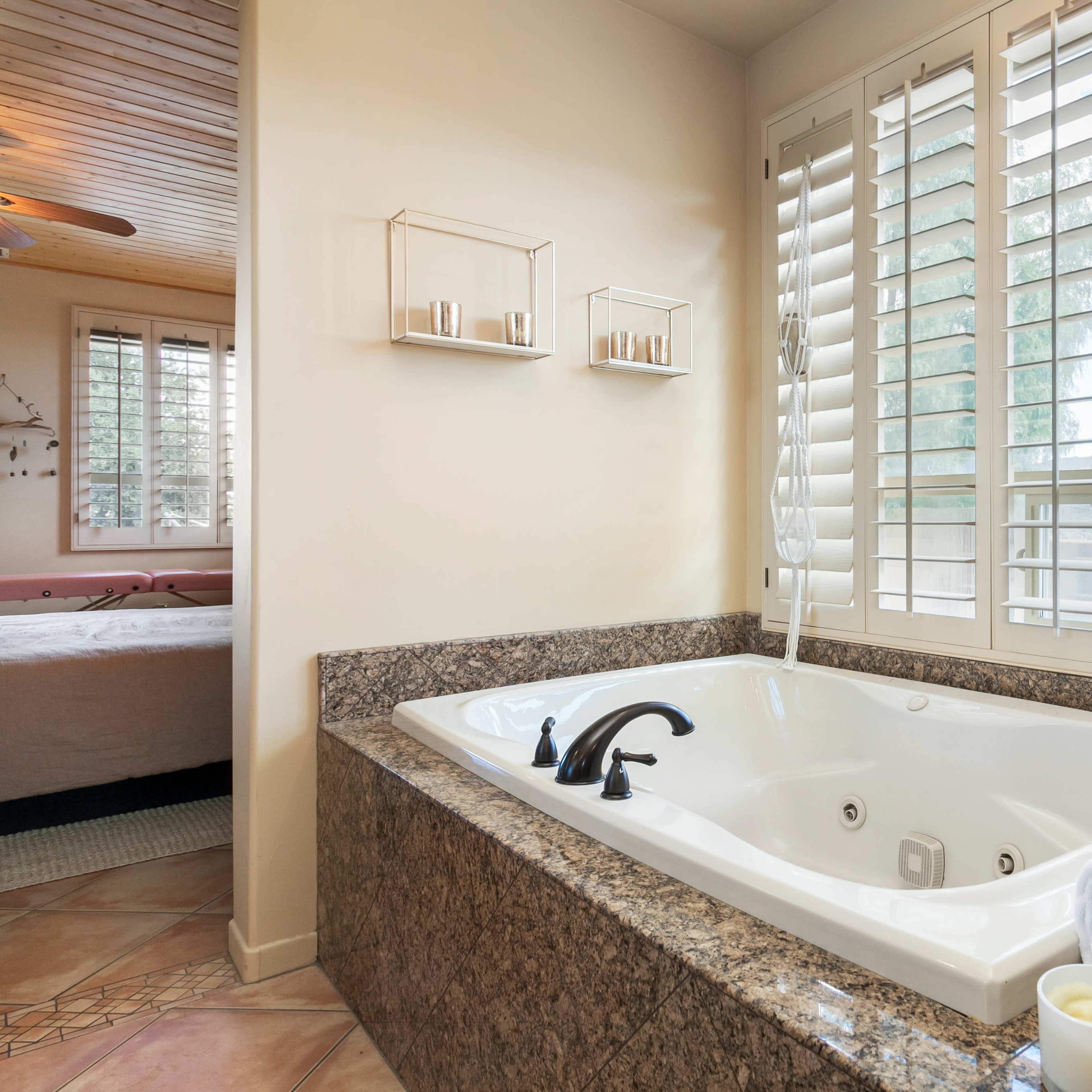A bathroom featuring a large jetted bathtub with a granite surround, beside a window covered with plantation shutters.
